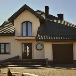 Elewacja domu jednorodzinnego z ciemną stolarką okienną i drzwiową, ciemną bramą garażową segmentową i schodami wejściowymi wyłożonymi kostką.