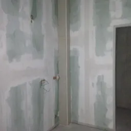 Ściana w trakcie prac przygotowawczych do wykończenia: płyty gipsowo-kartonowe, szpachlowanie w pasach, widoczne przewody elektryczne, surowy beton na podłodze, ościeżnica bez drzwi.