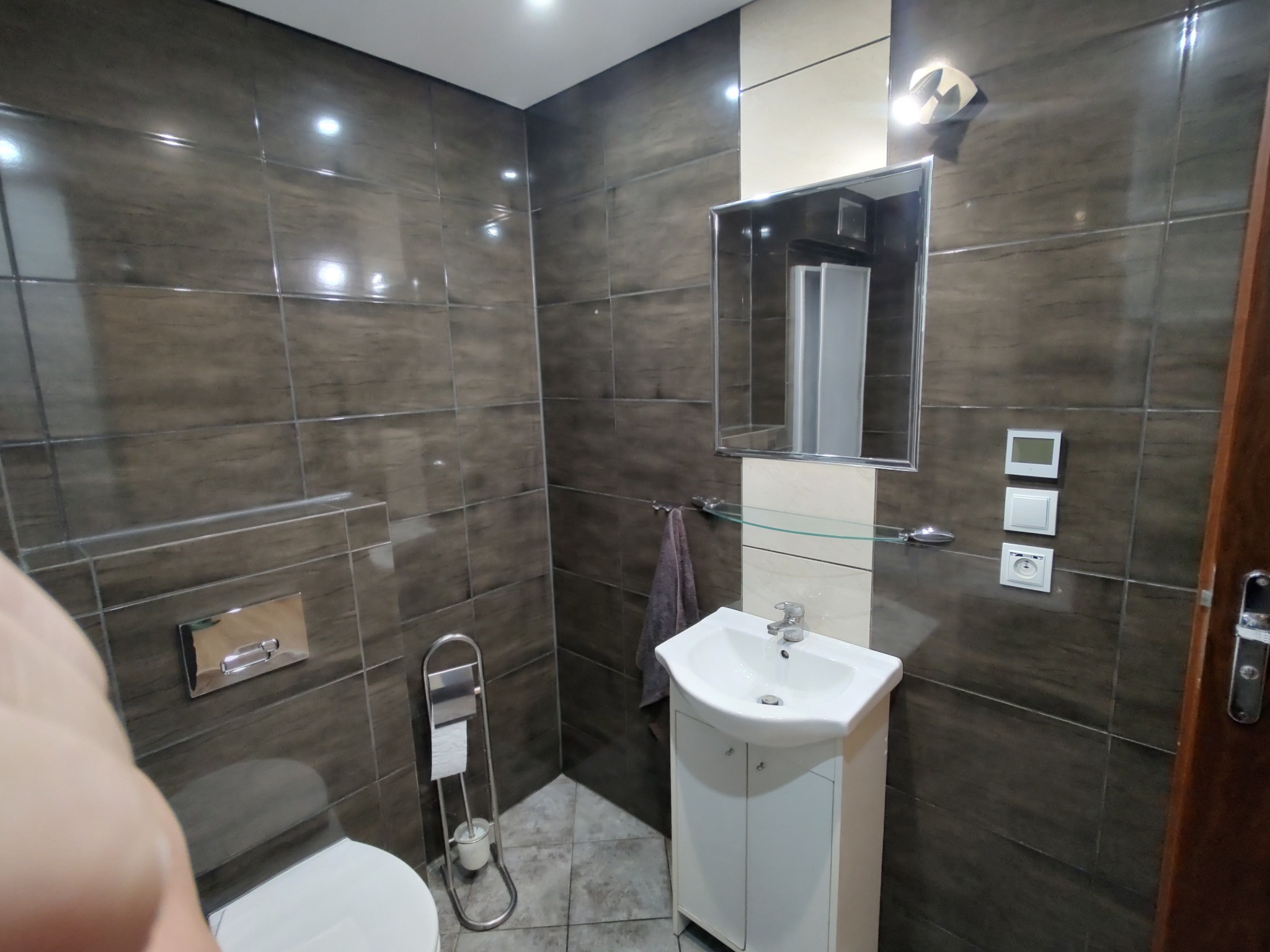 Wykończona łazienka z brązowymi płytkami, białą umywalką z szafką, lustrem i toaletą. Widoczne detale: włączniki, uchwyt na ręcznik i akcesoria toaletowe.