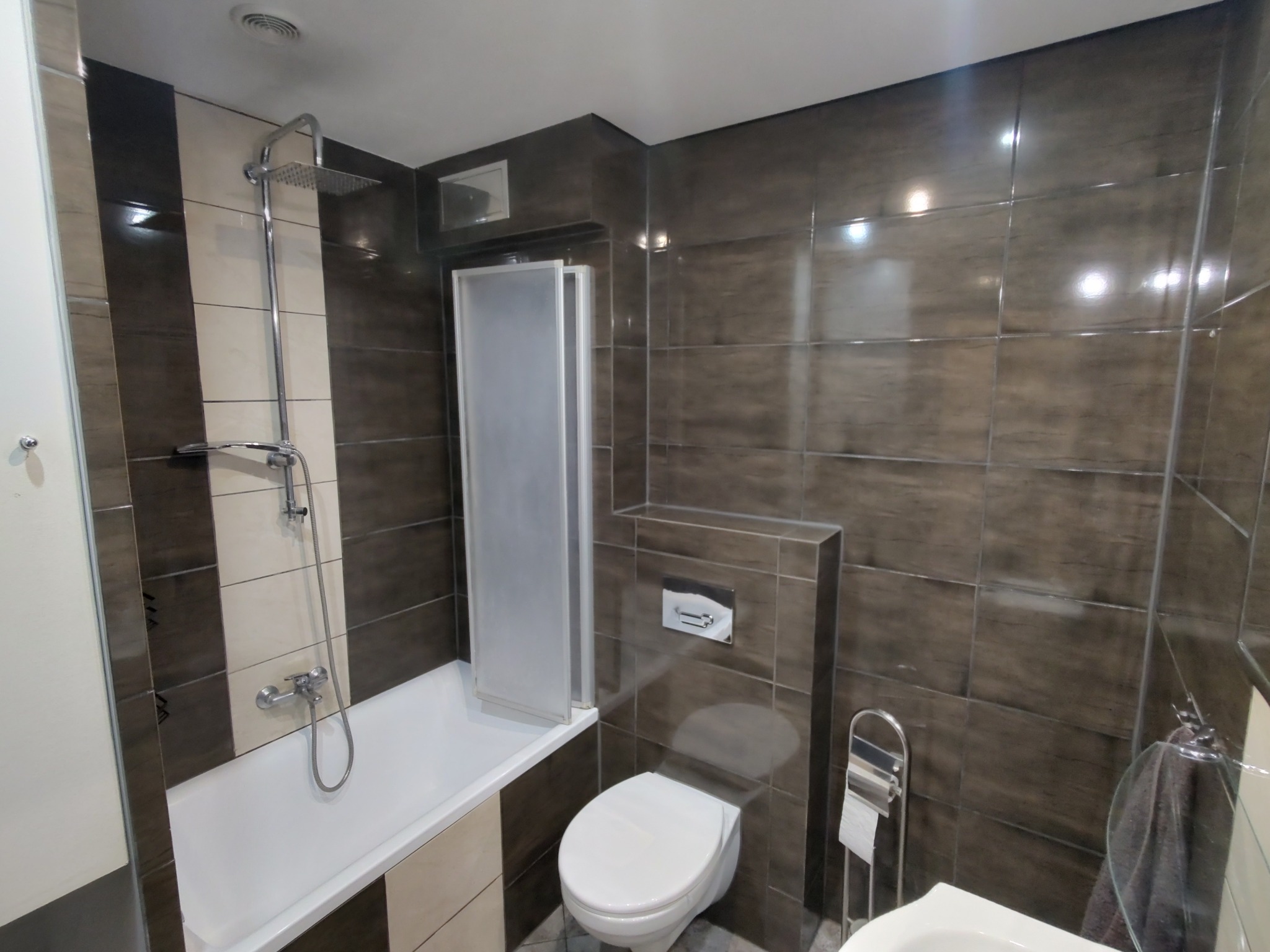 Wykończona łazienka z brązowymi płytkami, białą wanną z parawanem, toaletą i umywalką. Widoczne chromowane elementy armatury i akcesoriów łazienkowych.