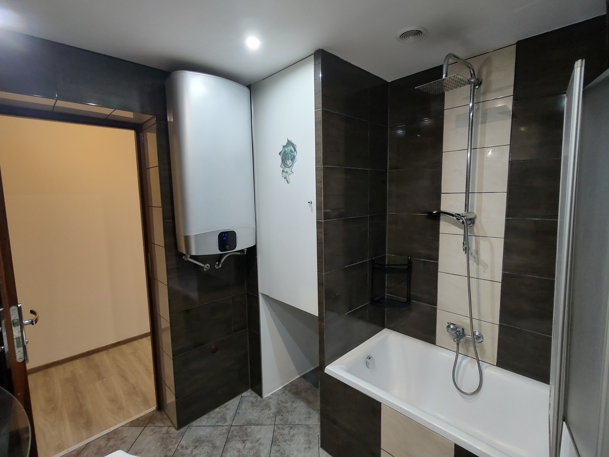 Nowoczesna łazienka po remoncie z brązowo-białymi płytkami, białą wanną i bojlerem. Widoczne detale wykończenia i oświetlenie.