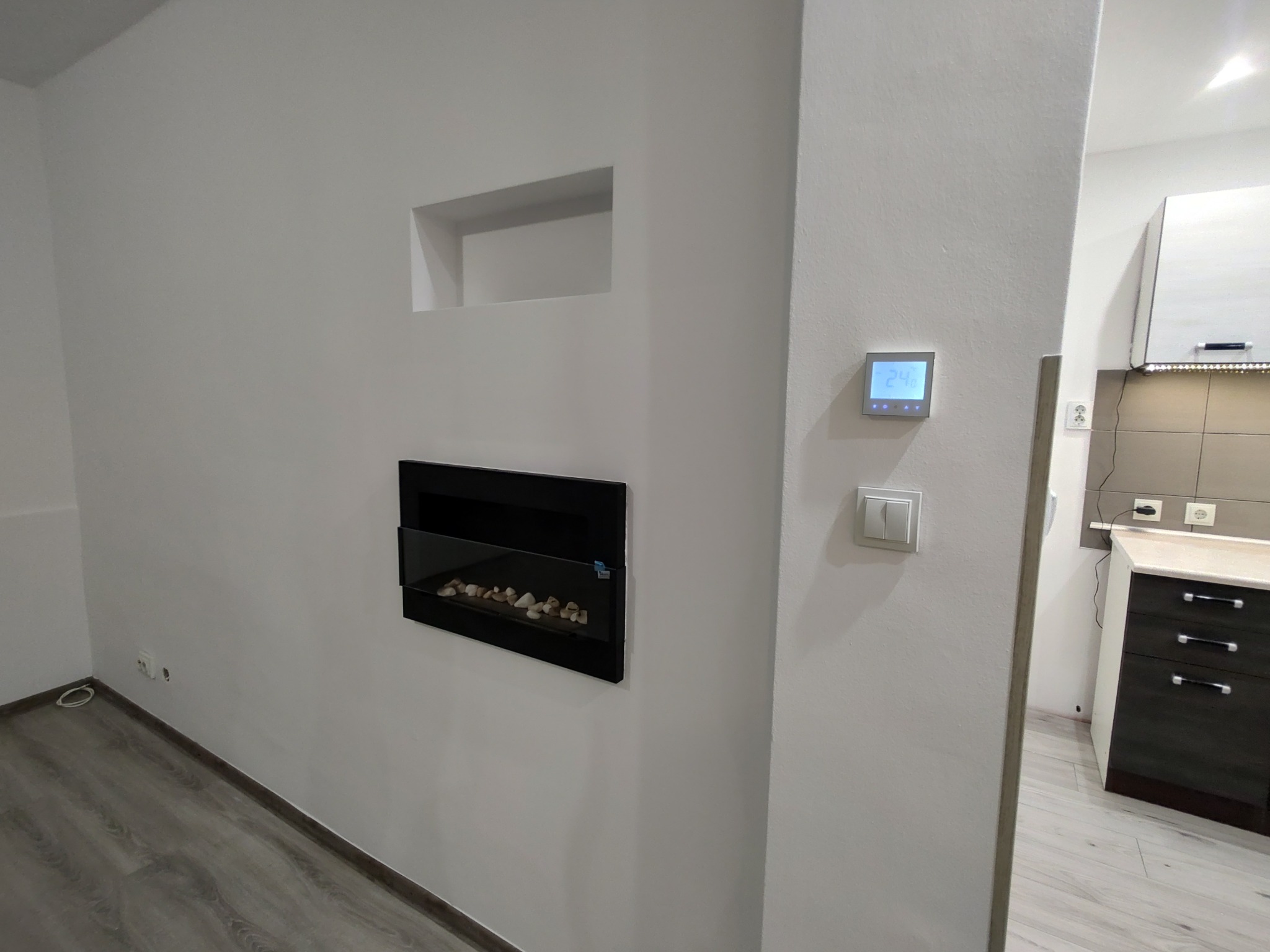 Minimalistyczne wnętrze po remoncie: białe ściany, panel podłogowy, wbudowany kominek elektryczny i widok na kuchnię z nowoczesną zabudową.