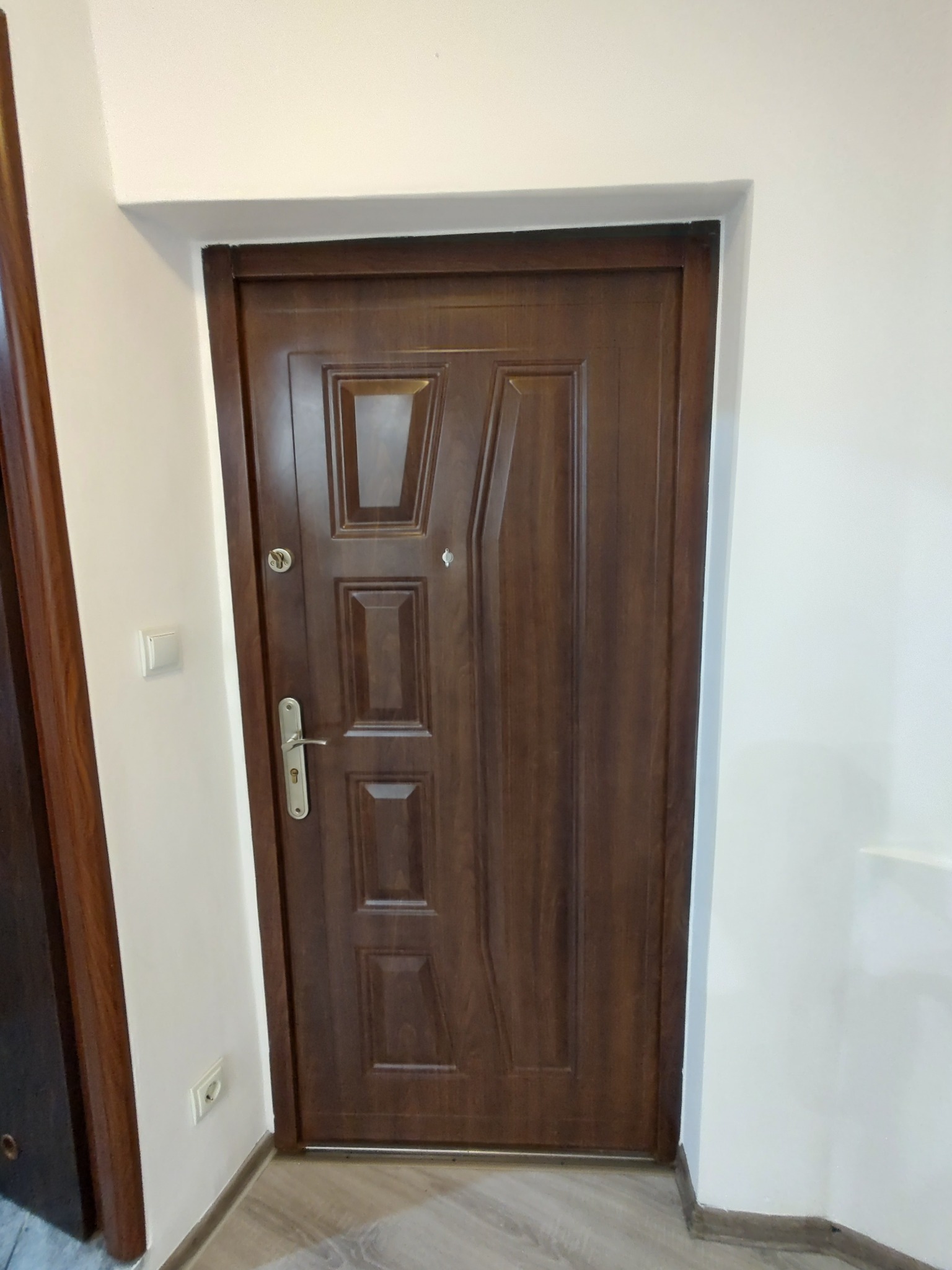 Ciemnobrązowe, nowoczesne drzwi wejściowe z panelem i wizjerem, zamontowane w białej futrynie. Widoczny fragment podłogi i ściany z włącznikiem.