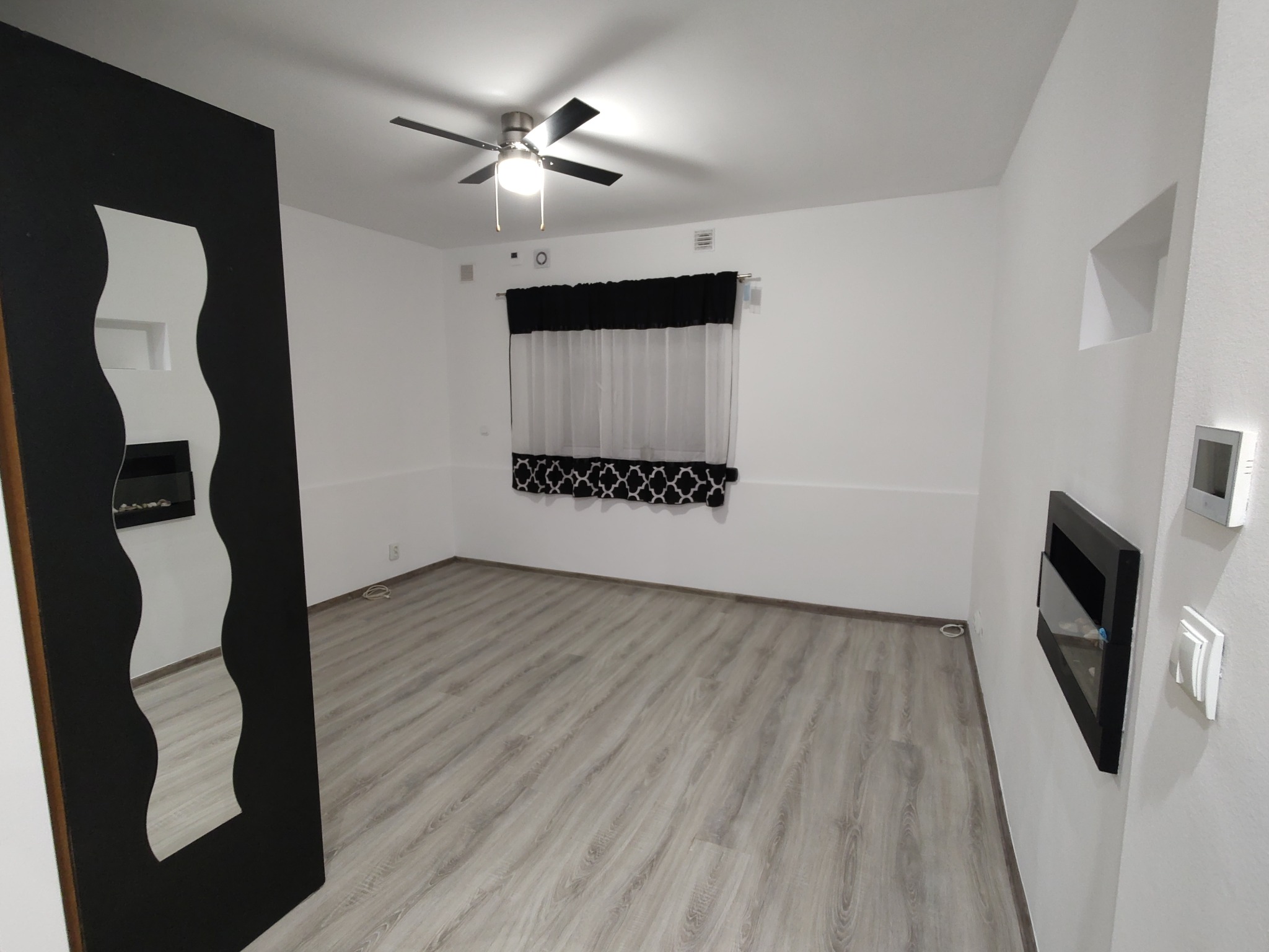 Jasne panele podłogowe w pokoju z białymi ścianami, czarnym kominkiem i lustrem o falistych krawędziach. Widoczny sufit z wentylatorem.