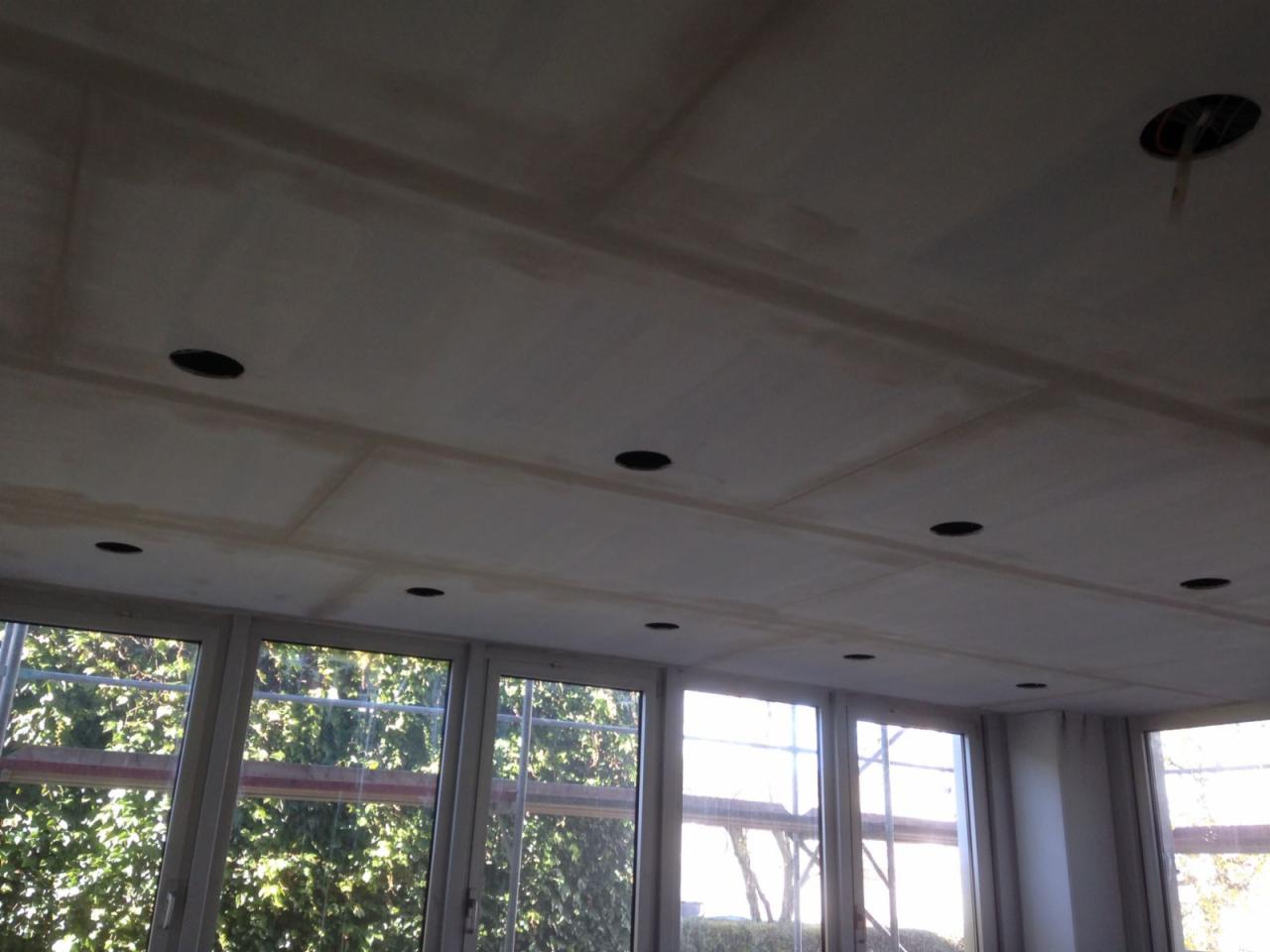 Sufit z płyt gipsowo-kartonowych po szpachlowaniu, z widocznymi otworami na oświetlenie punktowe, duże okna z widokiem na zieleń na zewnątrz.