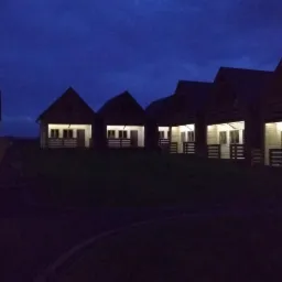 Rząd oświetlonych drewnianych domków z poddaszem w wieczornym oświetleniu, widoczny trawnik i droga.