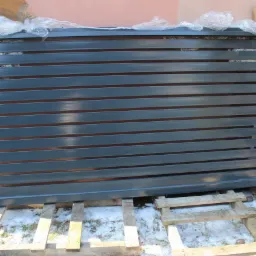 Szara metalowa brama lamelowa, zabezpieczona folią, leży na drewnianych paletach na śniegu, gotowa do montażu.