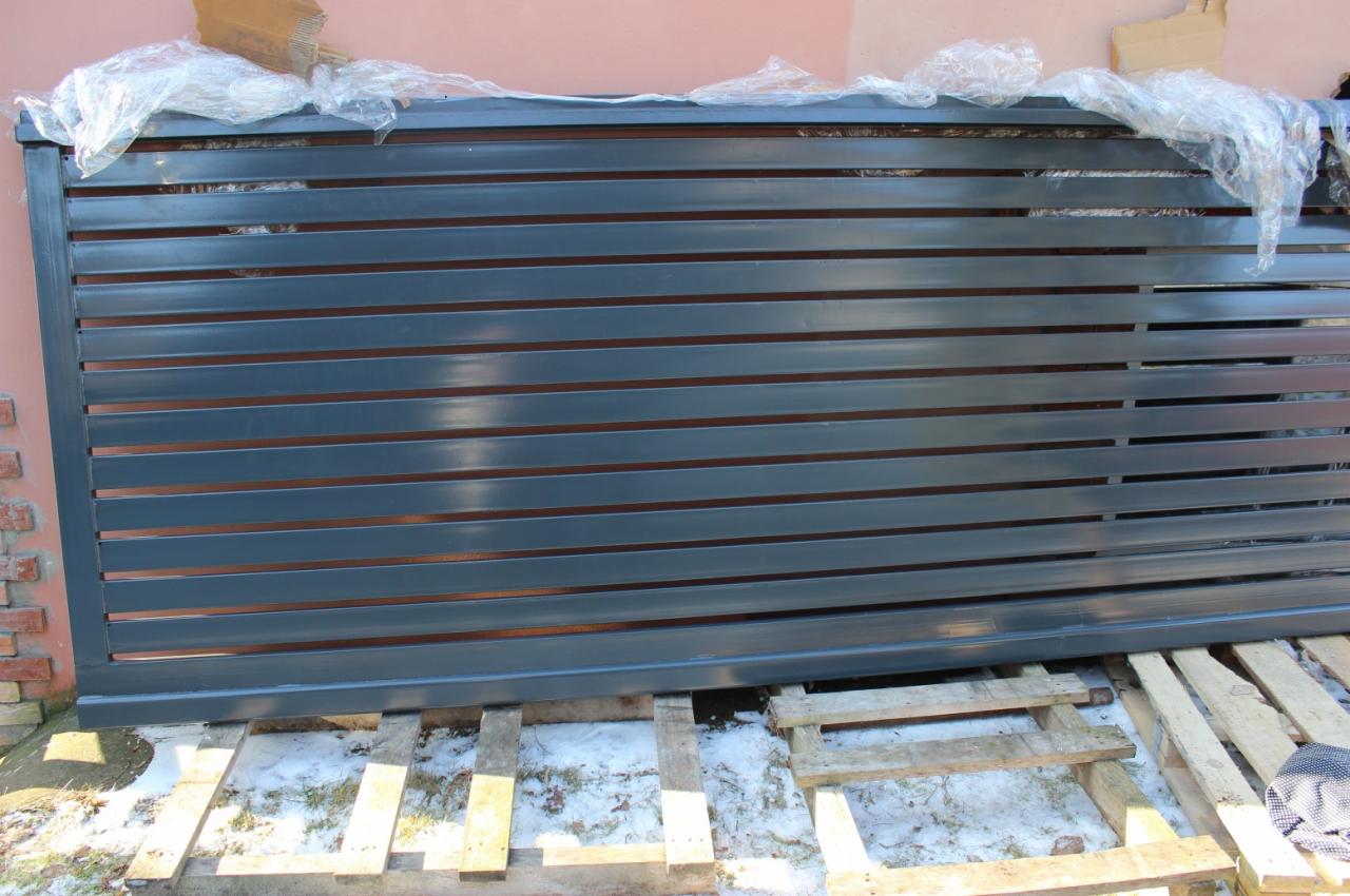 Szara metalowa brama lamelowa, zabezpieczona folią, leży na drewnianych paletach na śniegu, gotowa do montażu.