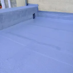 Jasnoniebieska, świeżo pomalowana płaska powierzchnia dachu z widocznym kominem wentylacyjnym i fragmentem rynny przy ścianie budynku.