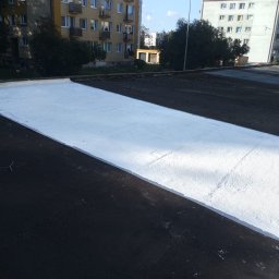 PHU FENIKS Robert Zmitrowicz - Biała powłoka hydroizolacyjna na czarnym dachu płaskim bloku mieszkalnego w słoneczny dzień. Częściowo pokryty dach z widoczną fakturą.