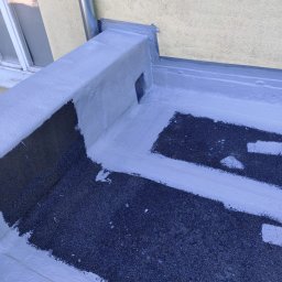 PHU FENIKS Robert Zmitrowicz - Szczegółowe ujęcie płaskiego dachu po hydroizolacji, z widoczną warstwą izolacyjną w kolorze szarym i fragmentem rynny spustowej. Widoczna faktura pokrycia dachowego.