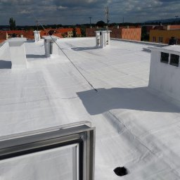 PHU FENIKS Robert Zmitrowicz - Biała, płaska powierzchnia dachu po renowacji z kominami wentylacyjnymi, widok z góry, na tle zabudowa miejska i pochmurne niebo. Widoczny fragment okna dachowego.