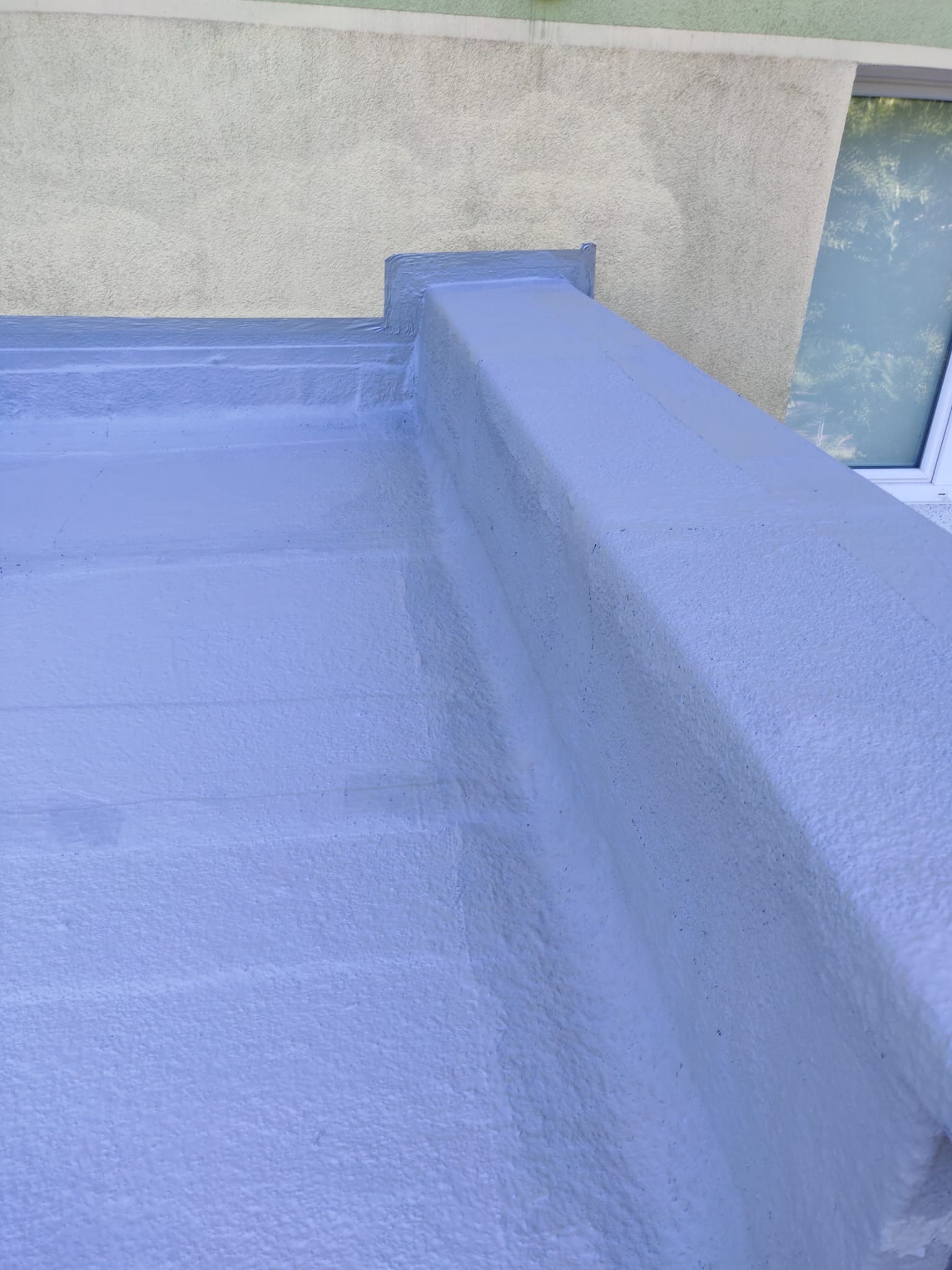 Pomalowany na szaro płaski dach budynku z widocznym fragmentem ściany i oknem. Widoczne łączenia i faktura farby. Kąt ujęcia z dołu.