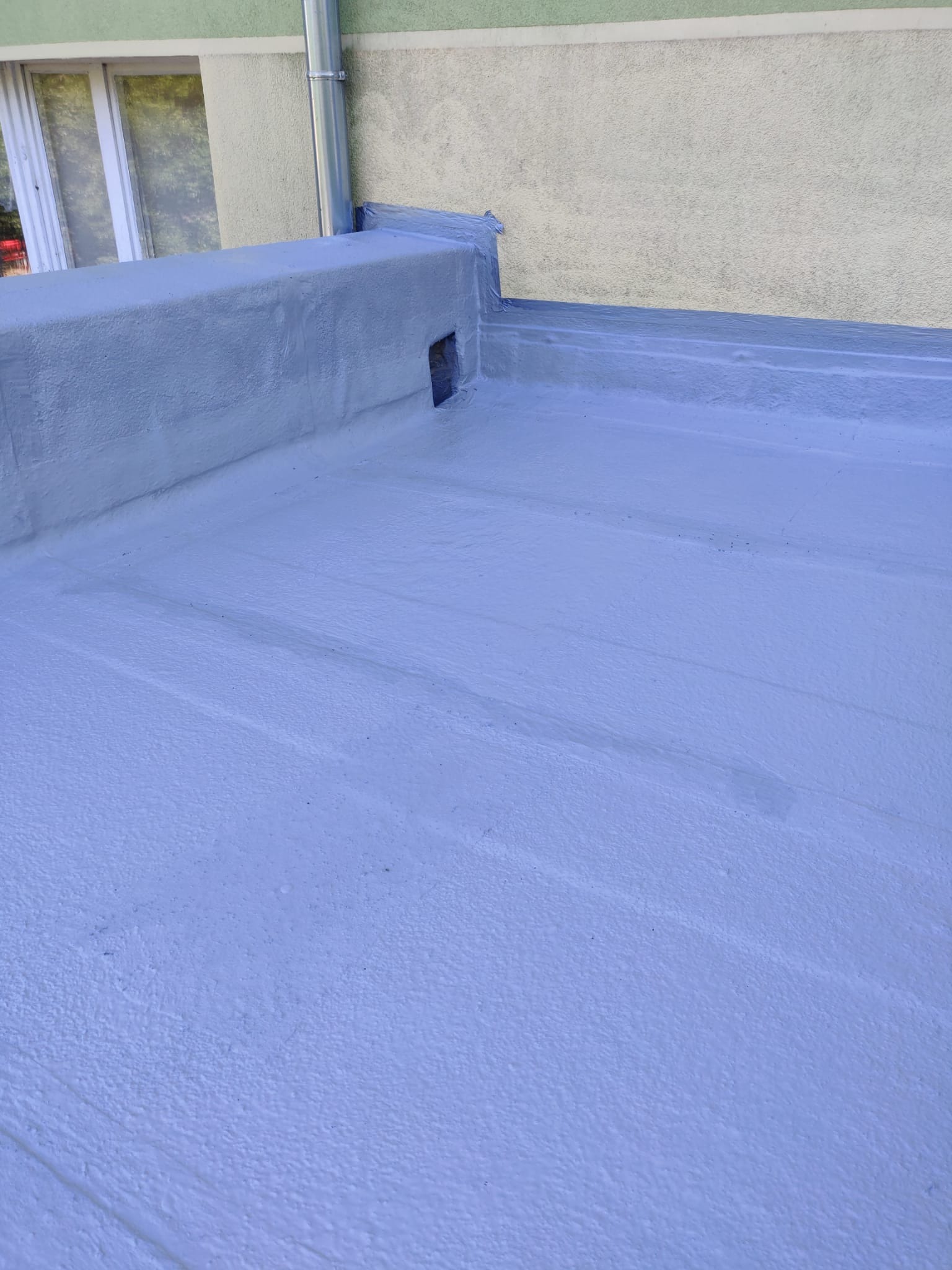 Jasnoniebieska, świeżo pomalowana płaska powierzchnia dachu z widocznym kominem wentylacyjnym i fragmentem rynny przy ścianie budynku.