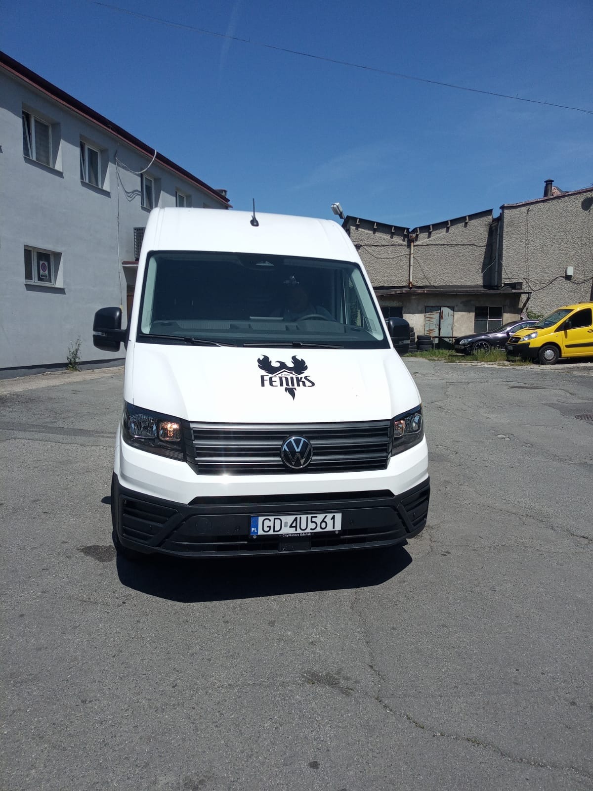 Biały samochód dostawczy z logo firmy dekarskiej FENIKS na tle budynków. Tablica rejestracyjna GD 4U561.