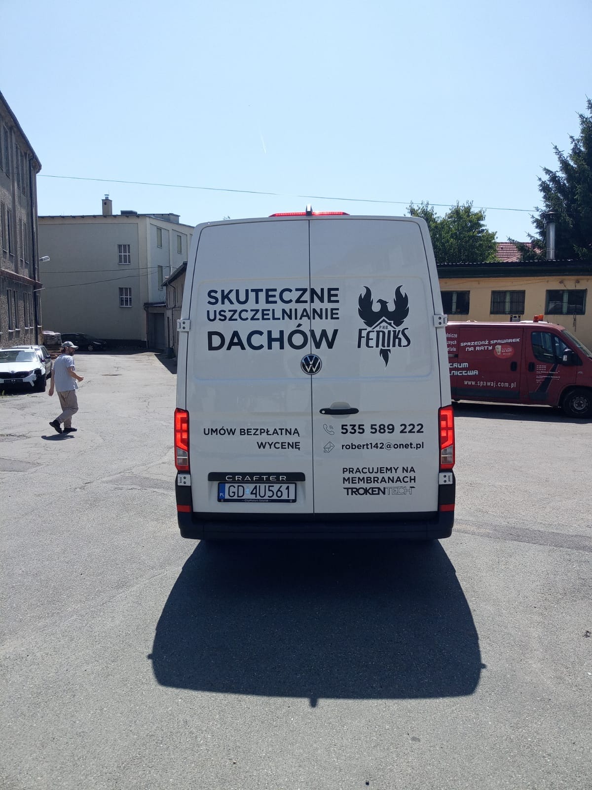 Biały samochód dostawczy z logo firmy 'Fenix' oferującej usługi uszczelniania dachów w Gdańsku. Widoczny numer telefonu i adres e-mail. Drugi samochód w tle.