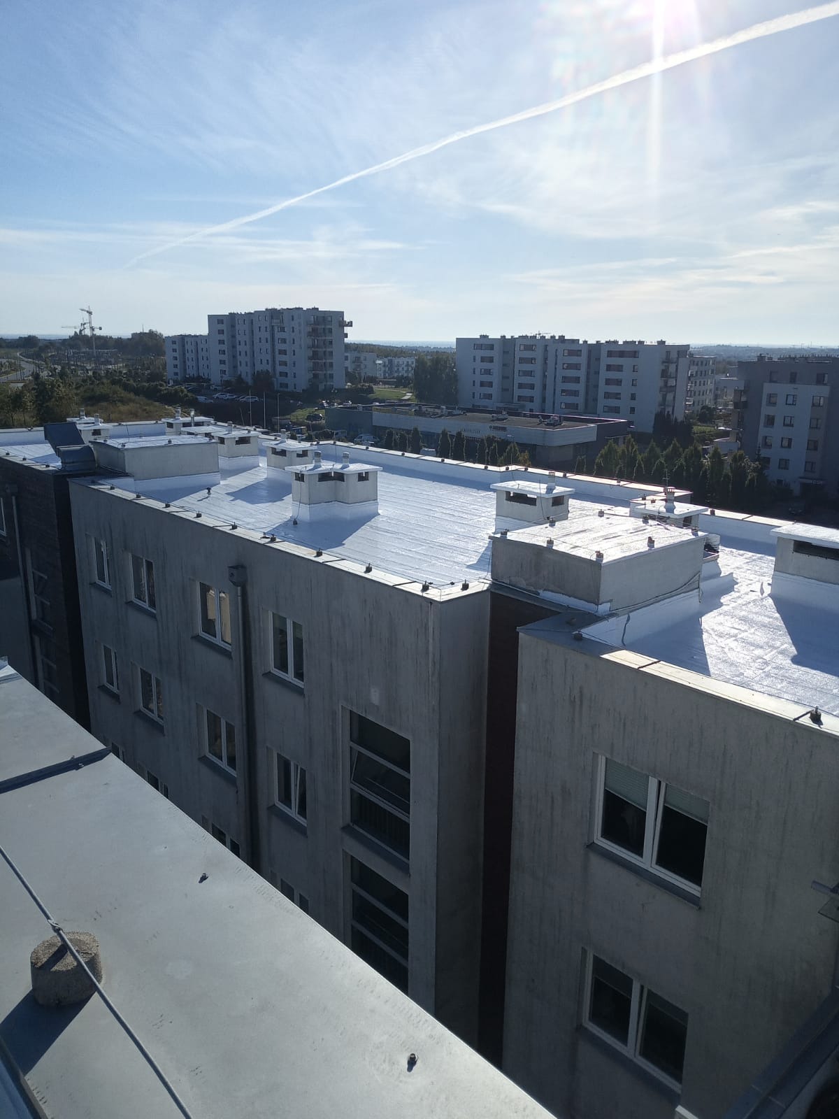 Białe, płaskie dachy bloków po renowacji w Gdańsku, widok z góry. Na tle błękitne niebo z delikatnymi chmurami i śladami po samolotach. Budynki mieszkalne w tle.