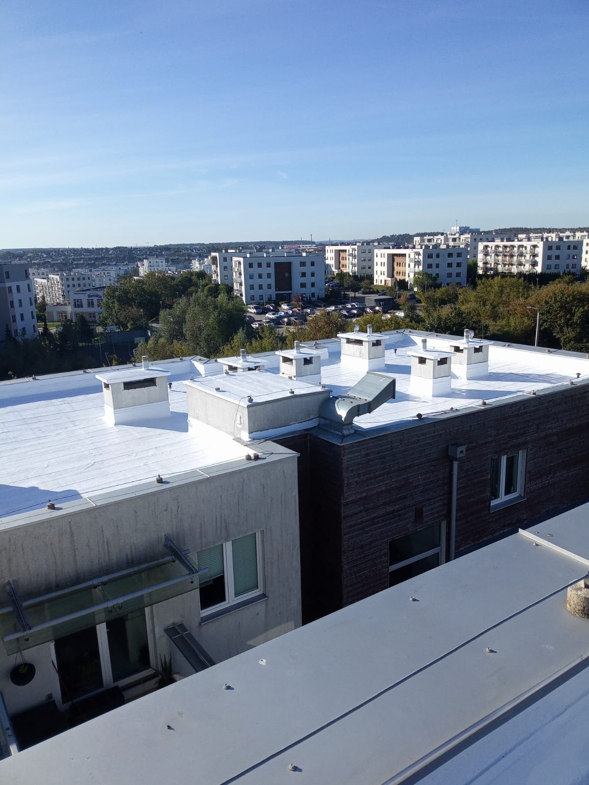 Biały dach z kominami na tle zabudowy miejskiej w słoneczny dzień. Widok z góry na płaski dach z elementami wentylacyjnymi i obróbkami blacharskimi.