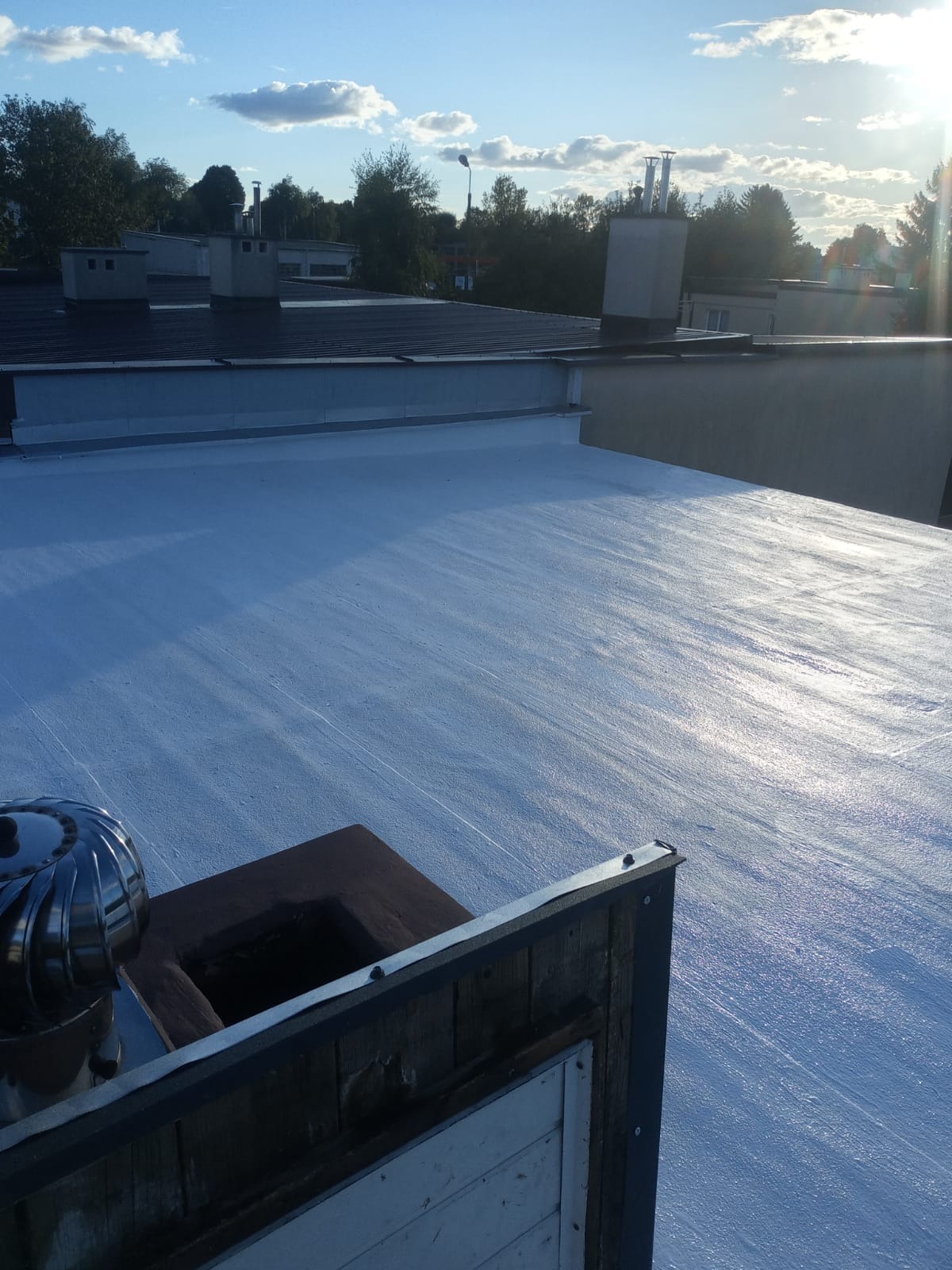 Biała, płaska powierzchnia dachu z kominem i turbiną w tle, widok z góry. Słoneczne oświetlenie podkreśla fakturę pokrycia. Elementy drewniane na pierwszym planie.