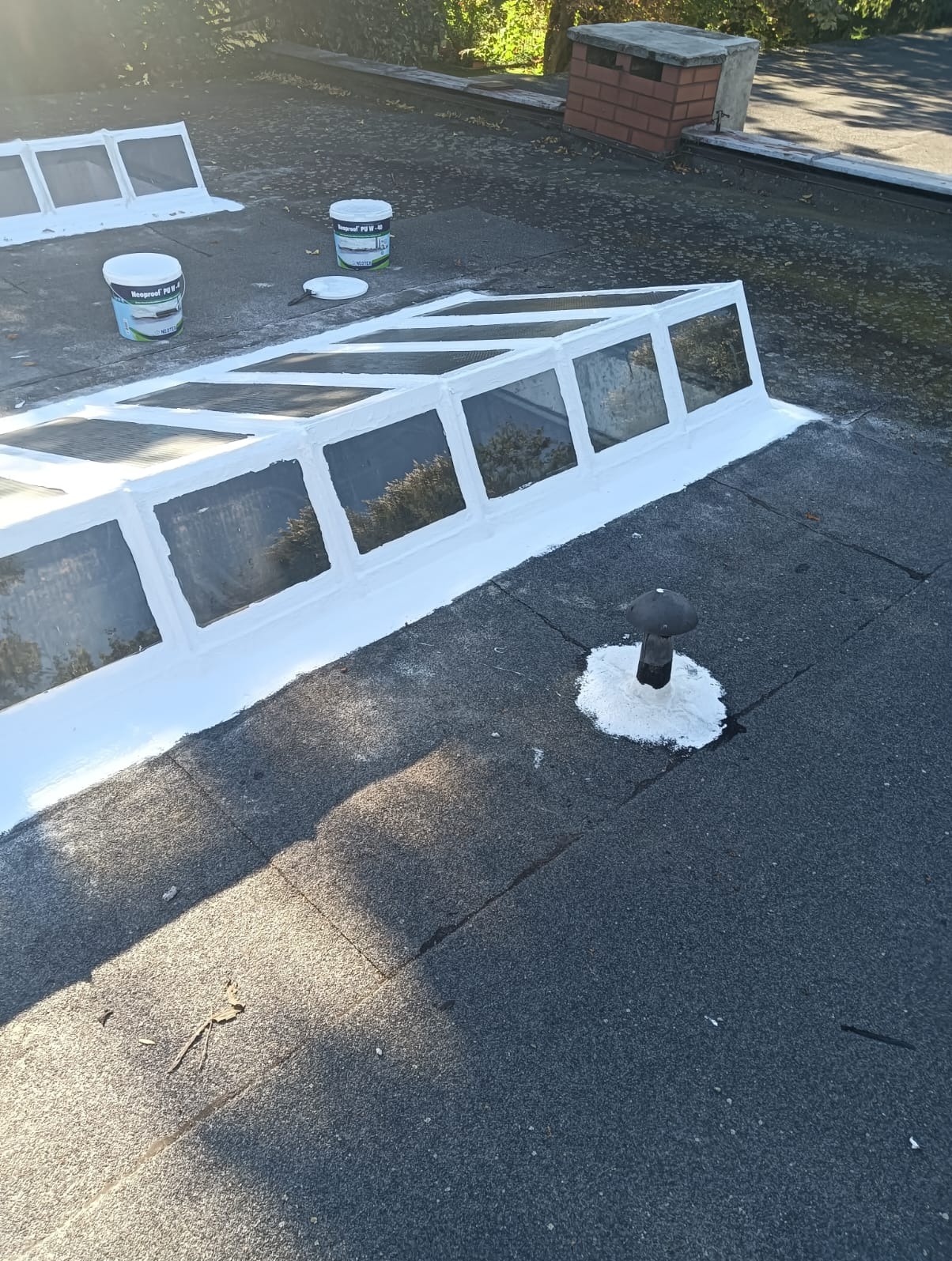 Płaski dach pokryty papą po uszczelnieniu świetlika dachowego i kominka wentylacyjnego białą masą uszczelniającą. Widoczne puszki z preparatem.