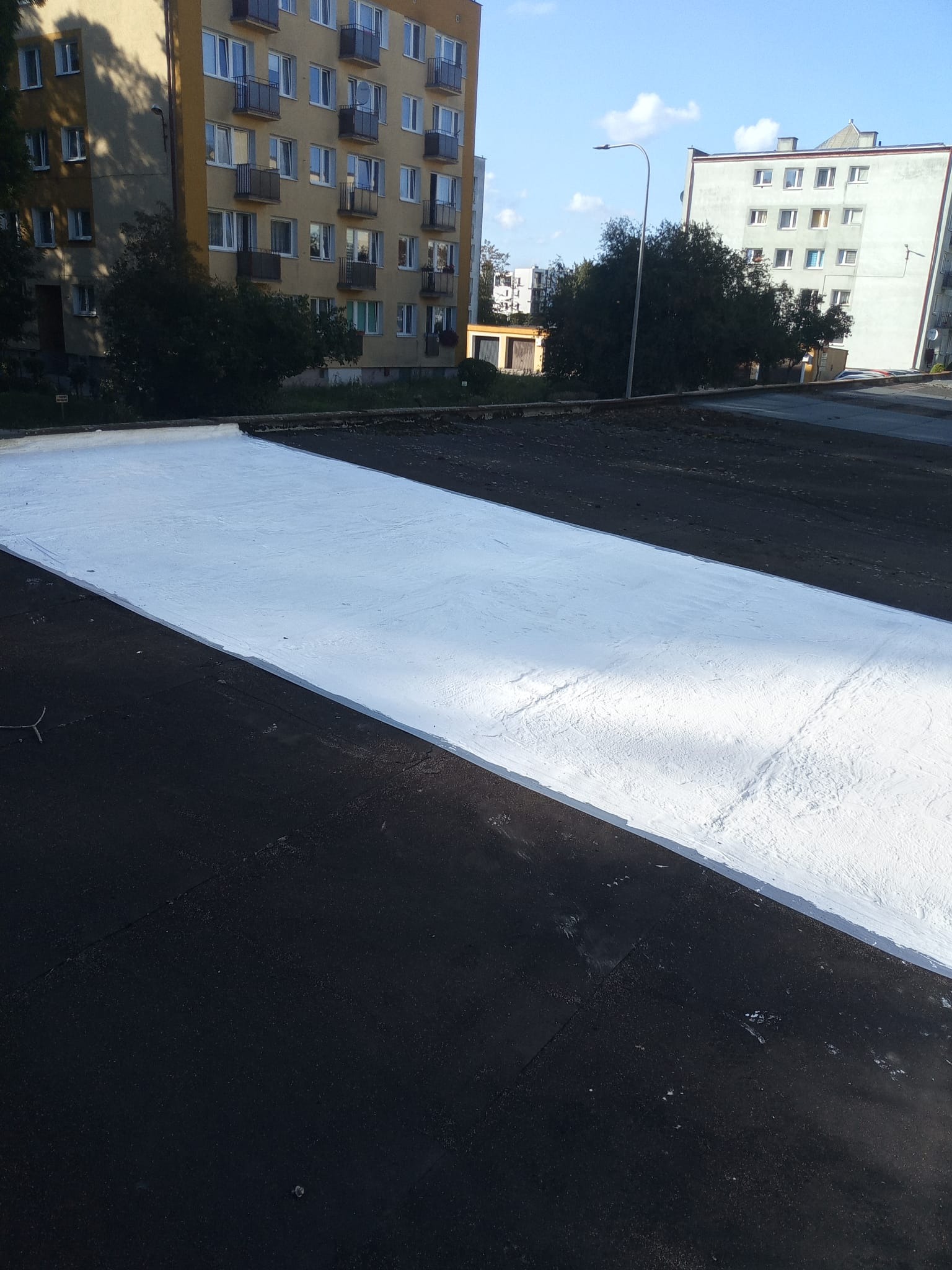 Biała powłoka hydroizolacyjna na czarnym dachu płaskim bloku mieszkalnego w słoneczny dzień. Częściowo pokryty dach z widoczną fakturą.