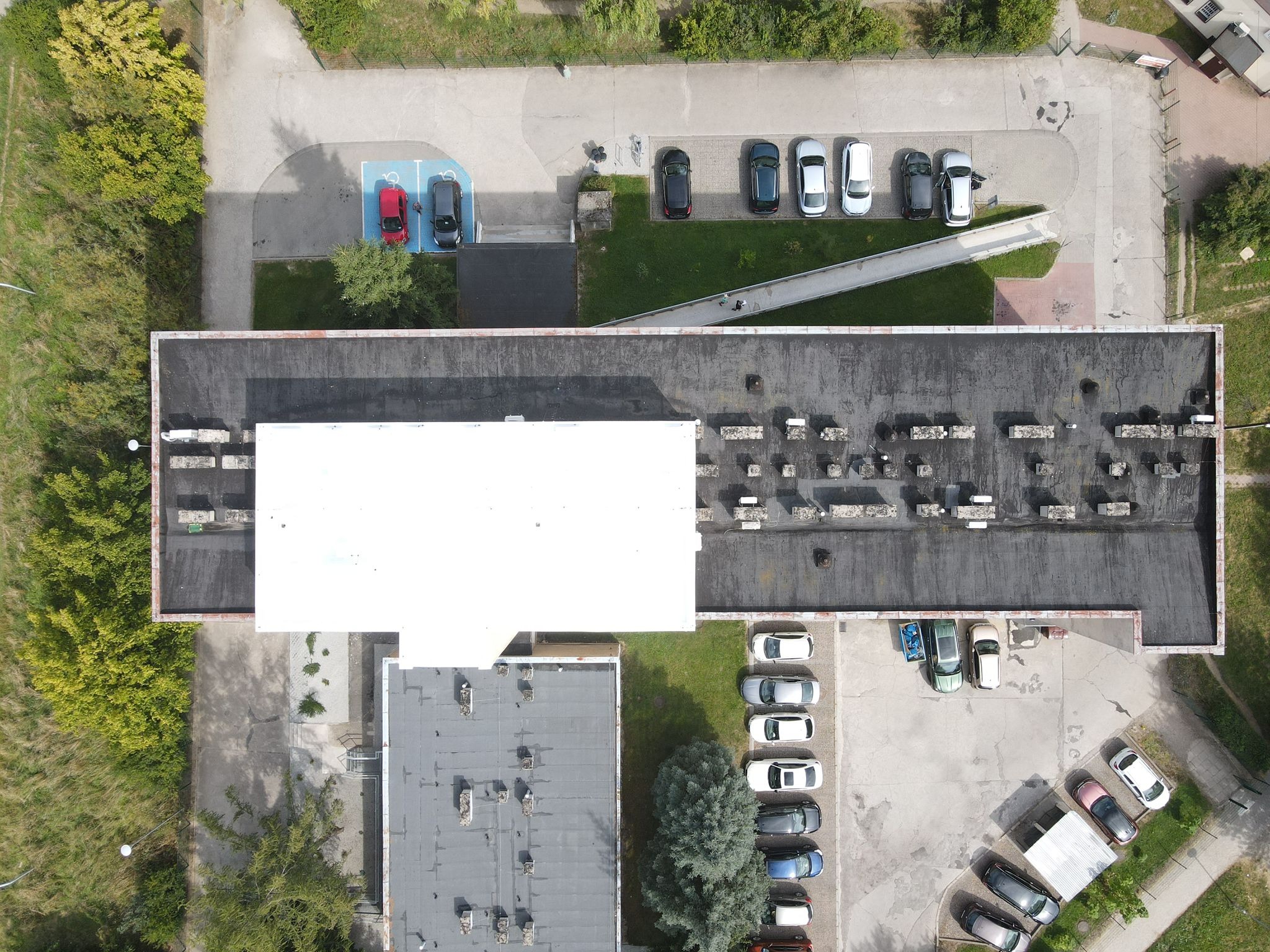 Widok z góry na płaski dach budynku z licznymi kominami wentylacyjnymi, otoczony parkingami i zielenią. Dach w odcieniach szarości, z białą, prostokątną nadbudówką.