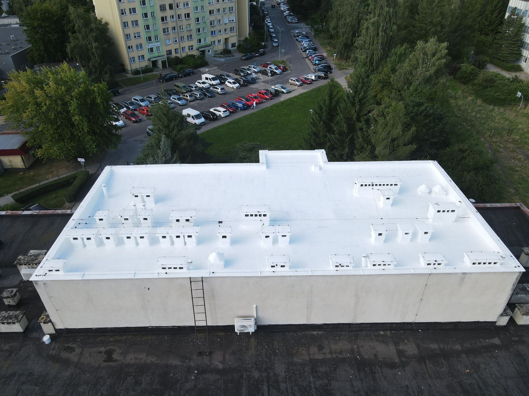Biały, płaski dach budynku z licznymi kominami wentylacyjnymi, widziany z góry. Obok dachu drabina i klimatyzator. W tle budynki mieszkalne i parking.