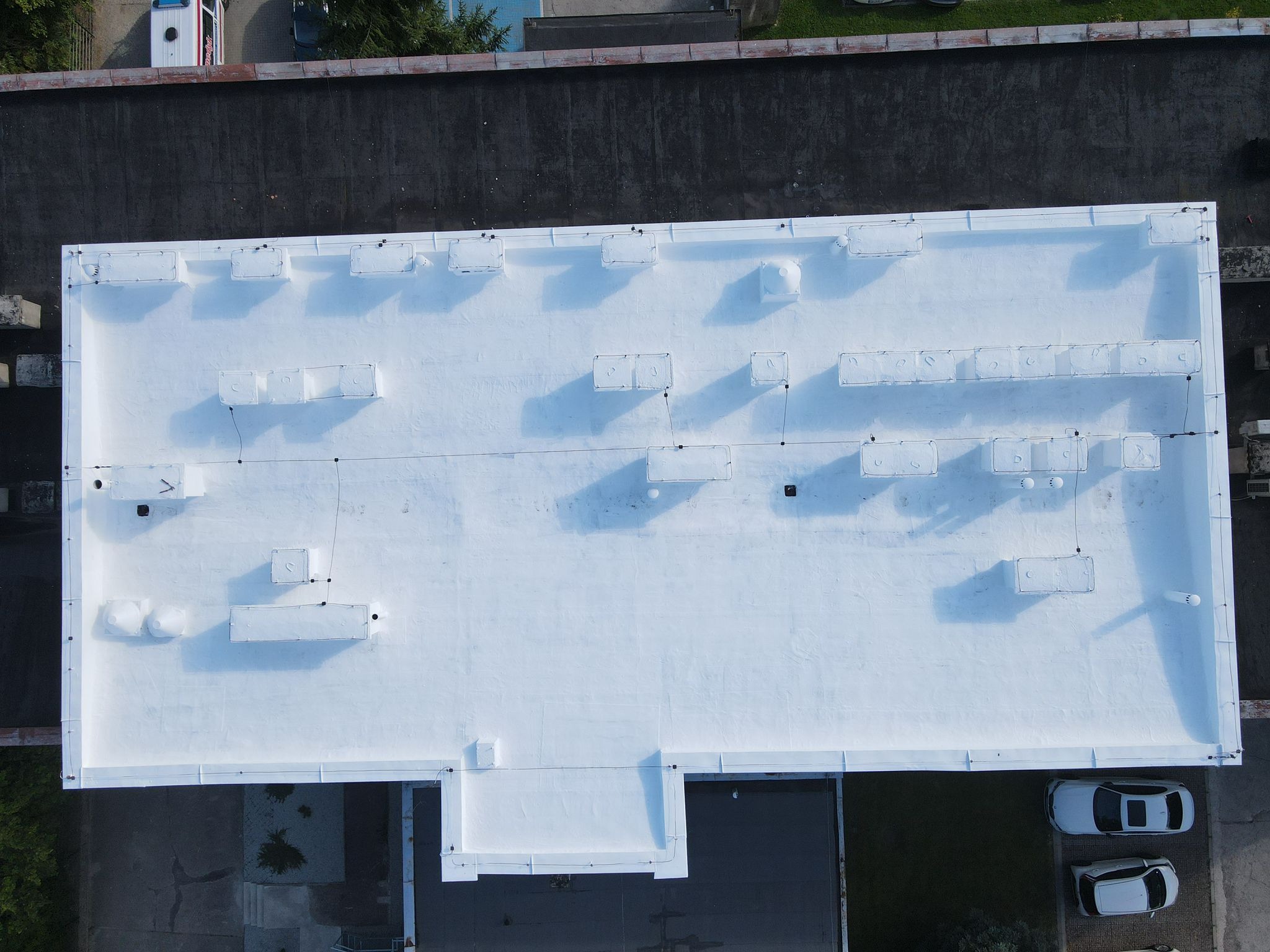 Biały, płaski dach budynku przemysłowego z licznymi świetlikami, widziany z góry. Obok dachu zaparkowane dwa samochody. W tle widoczny fragment innego dachu.