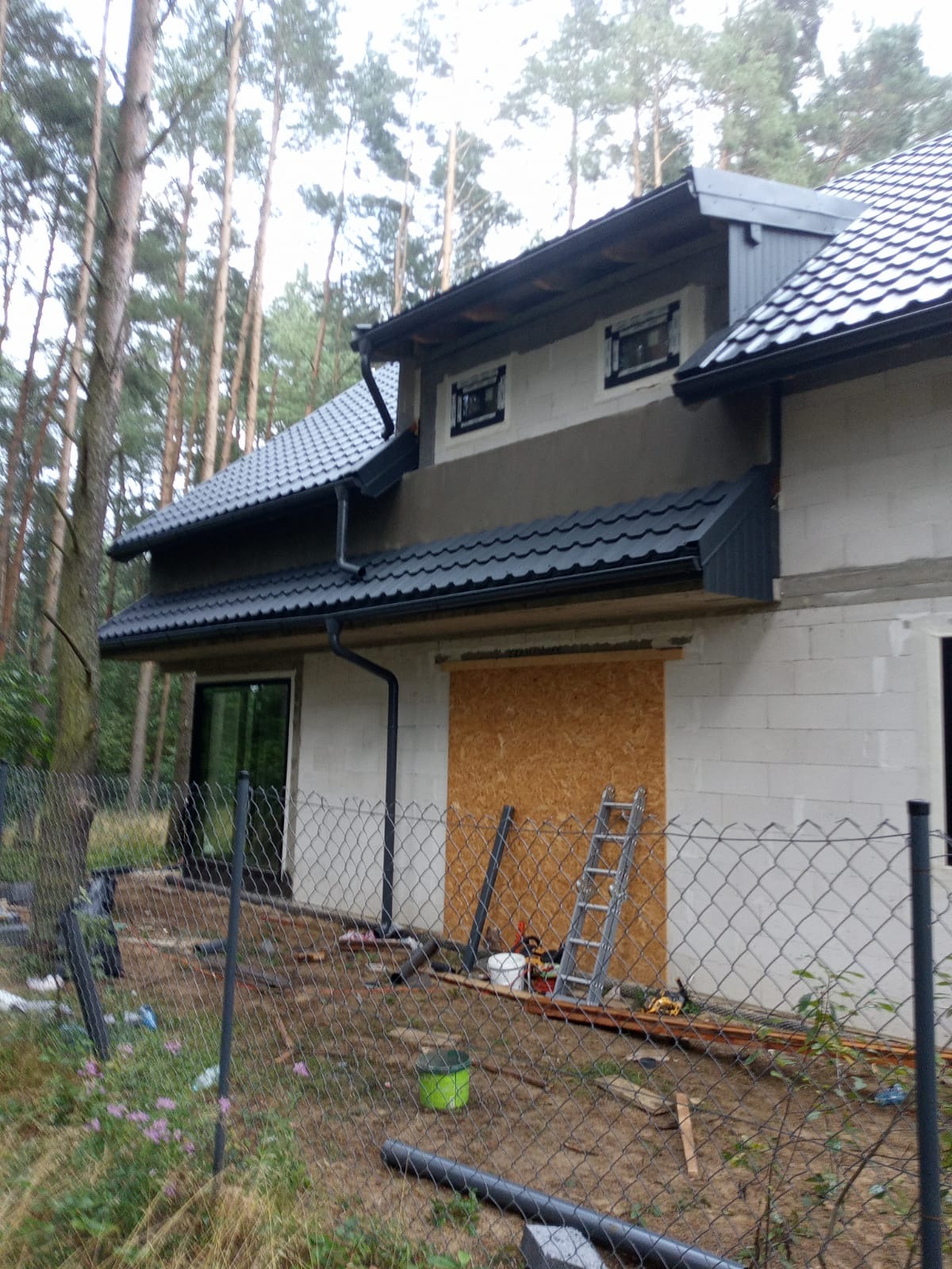 Dom w trakcie budowy z czarnym dachem z blachodachówki i rynnami. Część elewacji ocieplona, okna zamontowane. Widoczne narzędzia i drabina.