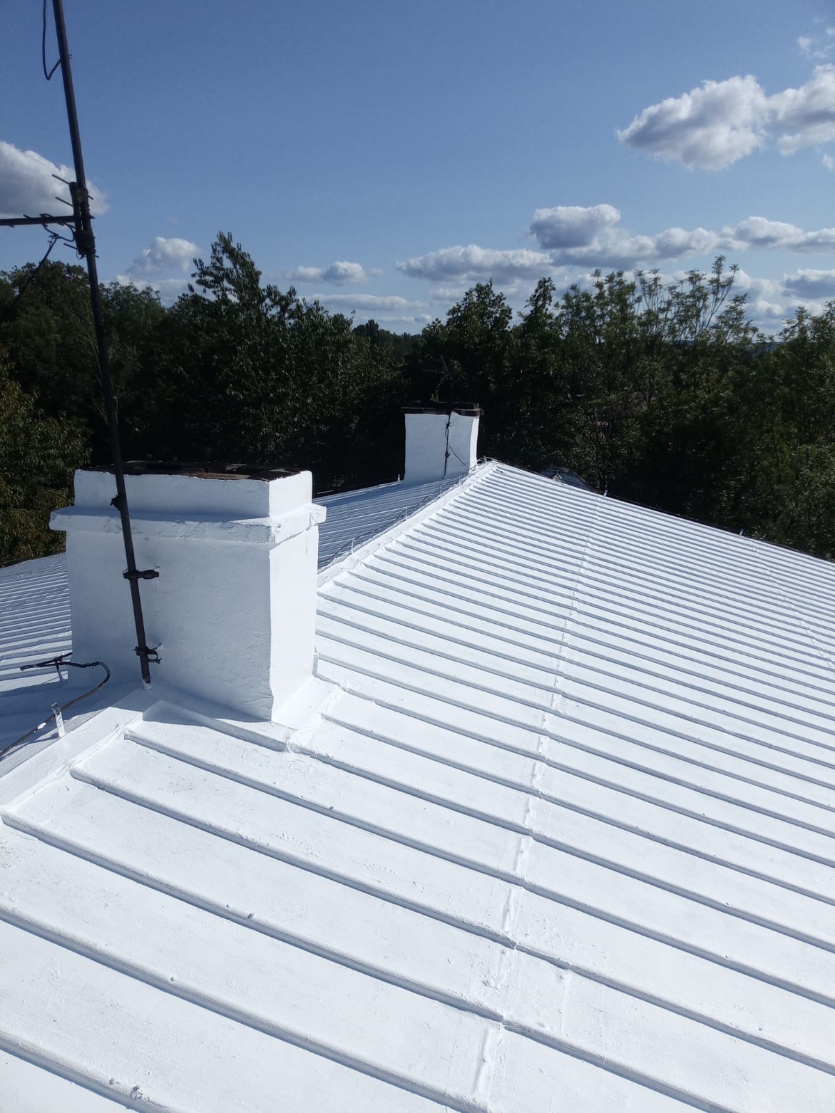 Biały dach z blachy trapezowej, dwa kominy pomalowane na biało, antena, widok z góry na tle zieleni i błękitnego nieba z chmurami.