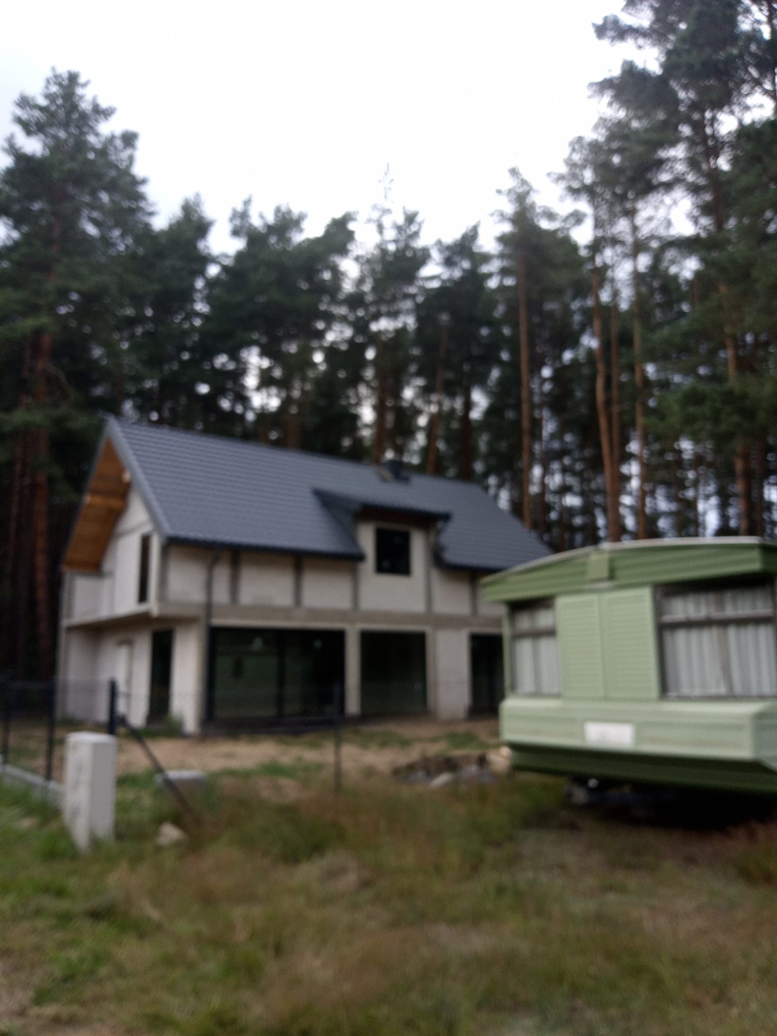 Surowy dom dwukondygnacyjny z ciemnym dachem w otoczeniu lasu, obok przyczepa kempingowa. Widok zza ogrodzenia.