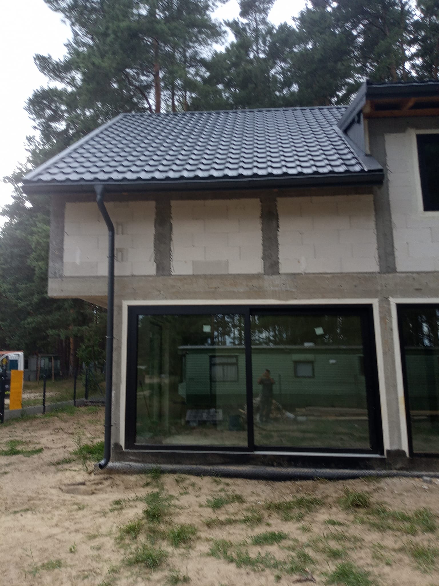 Nowoczesny dom w trakcie budowy z ciemnoszarą dachówką i czarnymi rynnami. Widoczne okna i surowy stan elewacji. Tło stanowią drzewa.