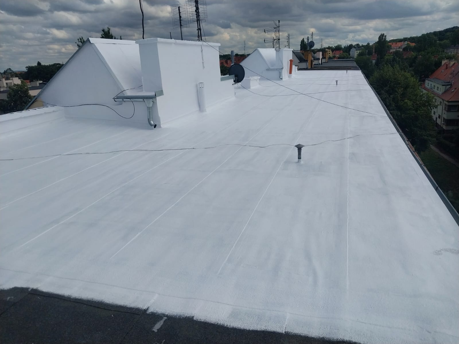 Biała, płaska powierzchnia dachu po renowacji, widok z góry. Widoczne kominy, anteny i elementy wentylacyjne. W tle zabudowa miejska i zieleń.