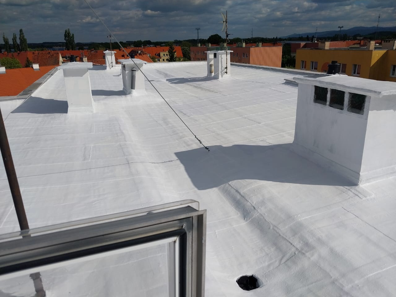 Biała, płaska powierzchnia dachu po renowacji z kominami wentylacyjnymi, widok z góry, na tle zabudowa miejska i pochmurne niebo. Widoczny fragment okna dachowego.