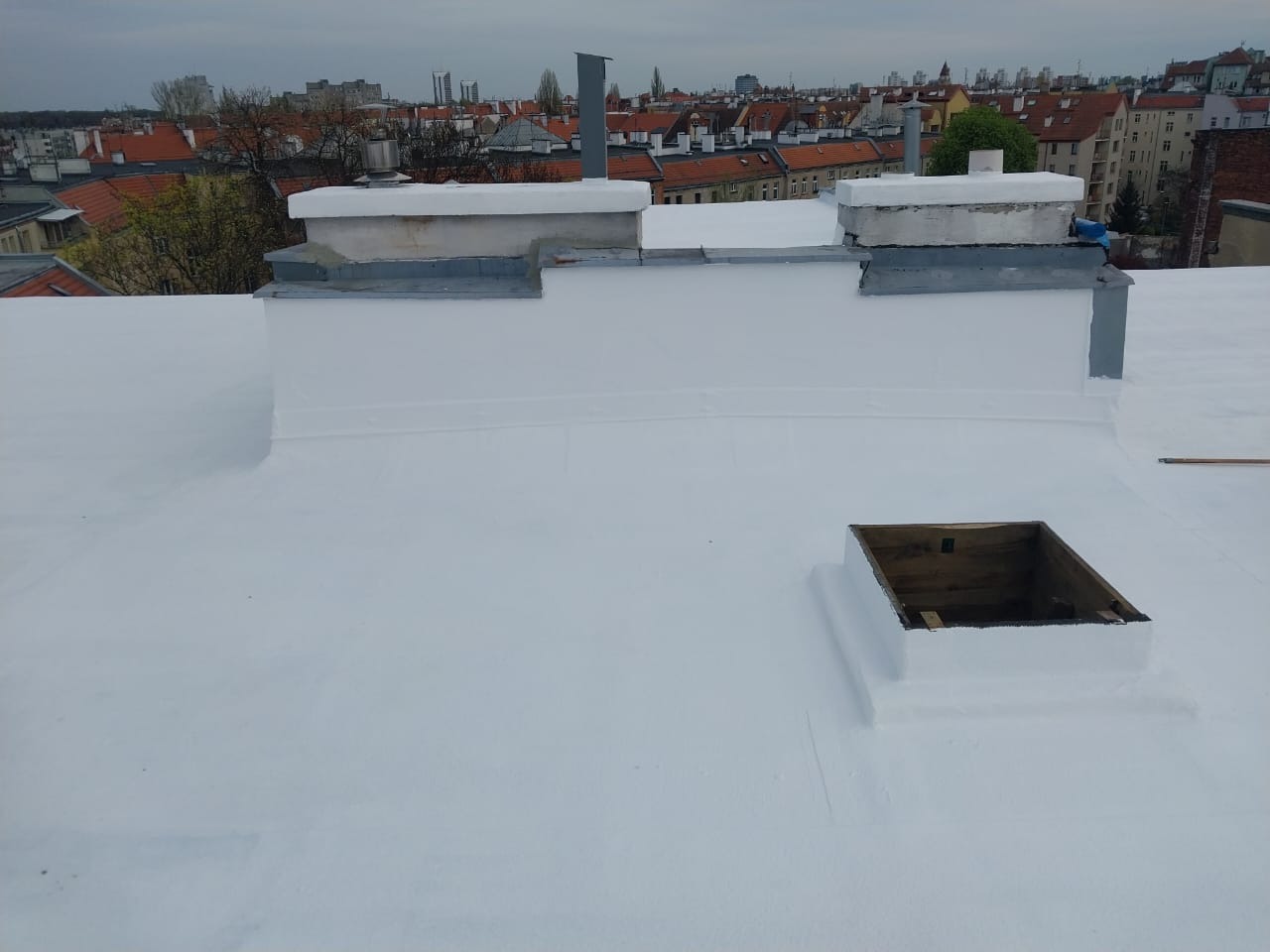Biała, płaska powierzchnia dachu po renowacji z kominami i otworem wentylacyjnym na tle zabudowy miejskiej w pochmurny dzień. Widoczne detale obróbek.