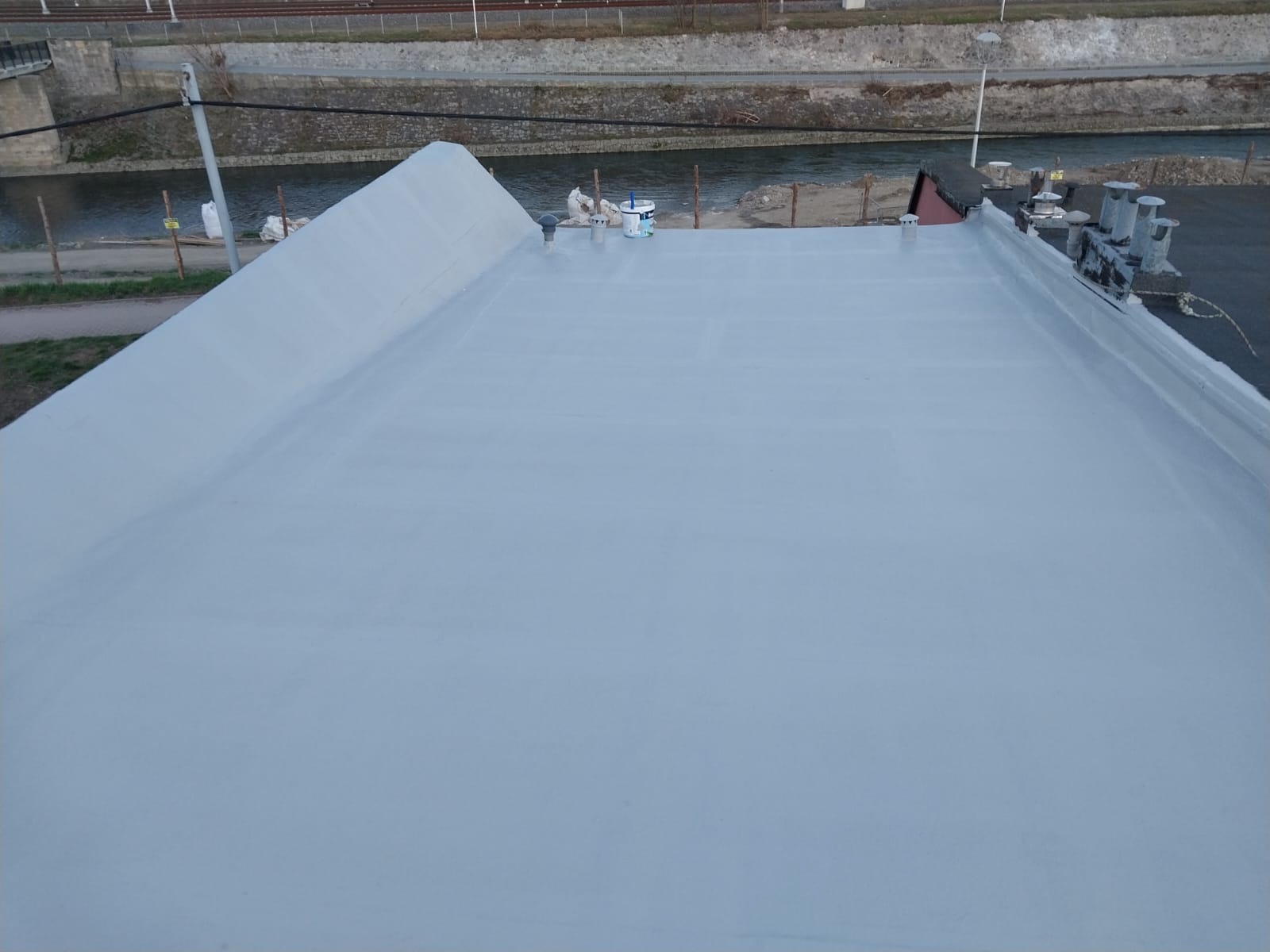 Gładka, szara powierzchnia dachu po renowacji, z widocznymi elementami wentylacyjnymi i puszką farby w tle, rzeka w oddali.