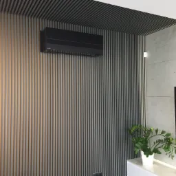 Czarny klimatyzator ścienny zamontowany na ścianie z pionowych paneli, w rogu pokoju z betonową ścianą i rośliną doniczkową na białej szafce.