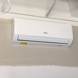 Klima Solution - Biały klimatyzator marki Gree zamontowany na ścianie z widocznym niedomalowaniem farby, pod sufitem podwieszanym z widocznymi kablami.