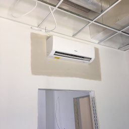 Klima Solution - Biały klimatyzator marki Gree zamontowany na ścianie w pomieszczeniu biurowym w trakcie wykańczania, widoczne instalacje pod sufitem podwieszanym i niedokończone malowanie ściany.