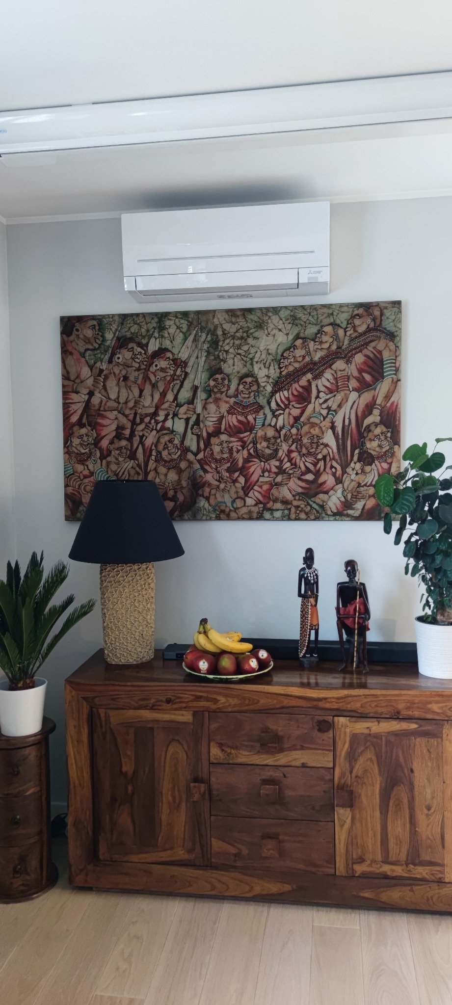 Biała jednostka klimatyzacyjna na ścianie nad drewnianą komodą z owocami, lampą i afrykańskimi figurkami, pod nią wisi obraz w etnicznym stylu.