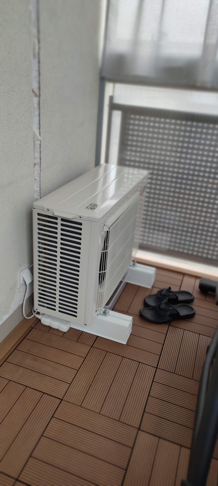 Biały klimatyzator zewnętrzny na balkonie z brązową podłogą z desek kompozytowych, widoczne czarne klapki i przedłużacz podłączony do gniazdka.