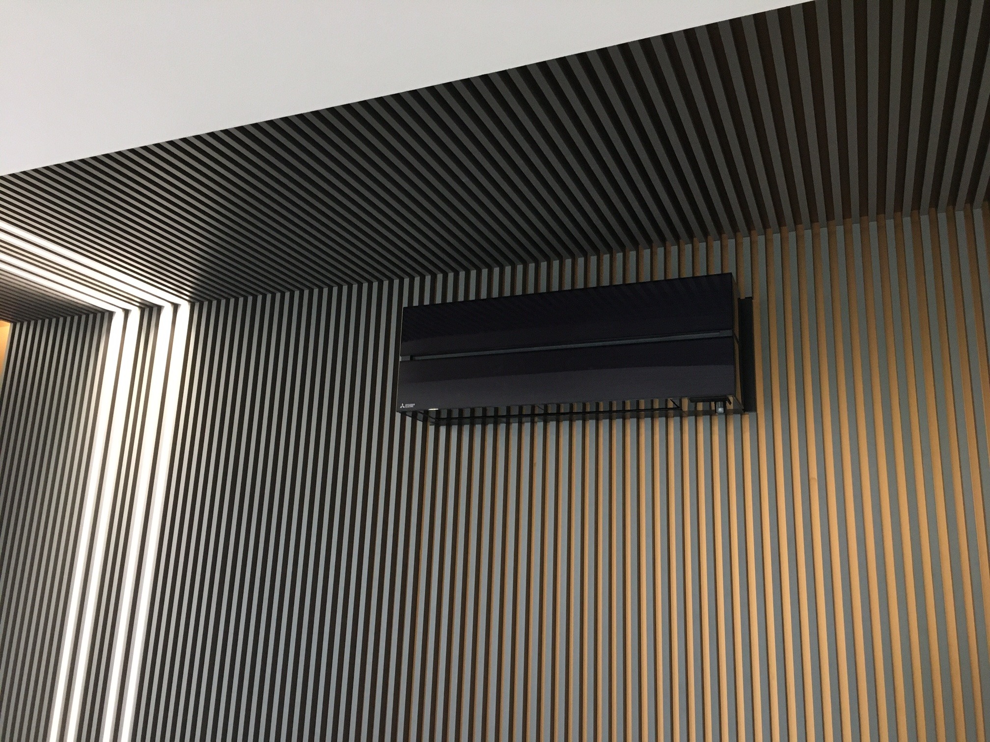 Czarny klimatyzator Mitsubishi Electric zamontowany na ścianie z pionowych, naprzemiennie ułożonych listew w kolorach szarości i ciepłego brązu, widok z dołu w nowoczesnym wnętrzu.