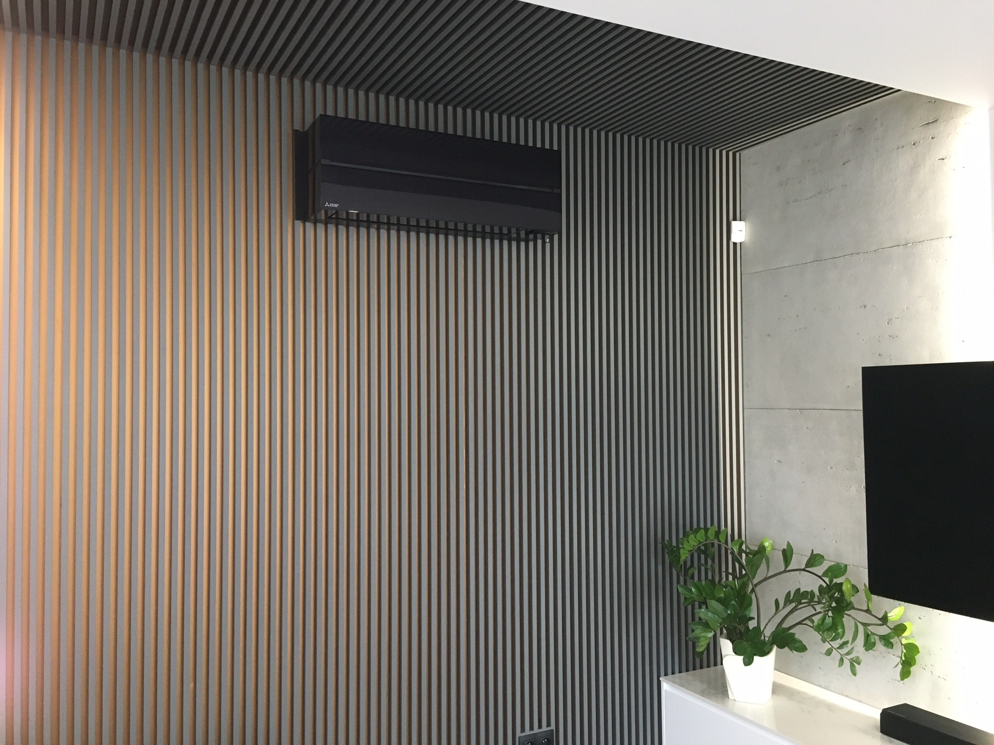 Czarny klimatyzator ścienny zamontowany na ścianie z pionowych paneli, w rogu pokoju z betonową ścianą i rośliną doniczkową na białej szafce.