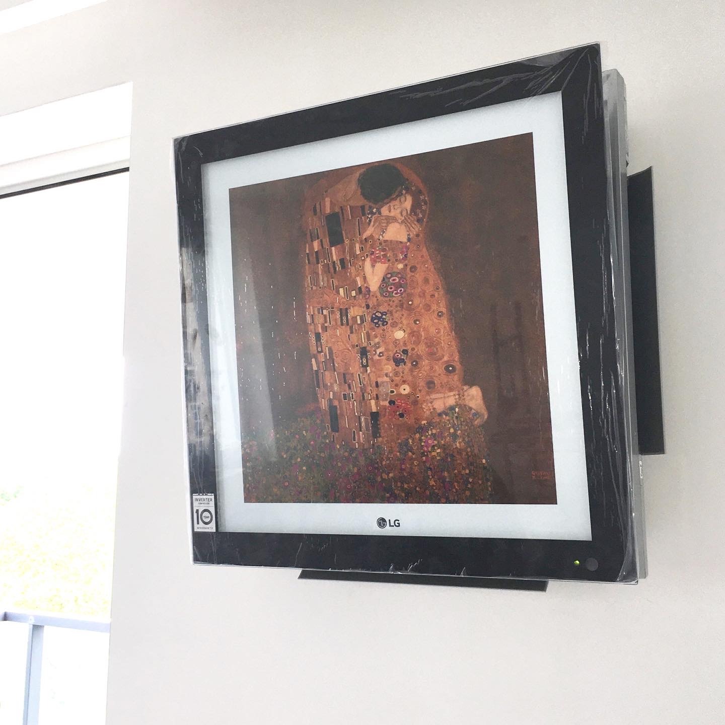 Klimatyzator LG Art Cool Gallery z obrazem 'Pocałunek' Gustava Klimta, jeszcze w folii ochronnej, zamontowany na białej ścianie.