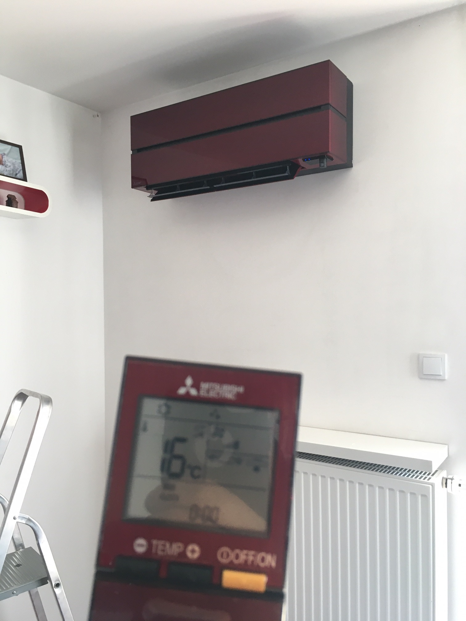 Bordowy klimatyzator Mitsubishi Electric zamontowany na białej ścianie w pomieszczeniu, z pilotem ustawionym na 16 stopni Celsjusza, widoczny fragment drabiny i grzejnika.