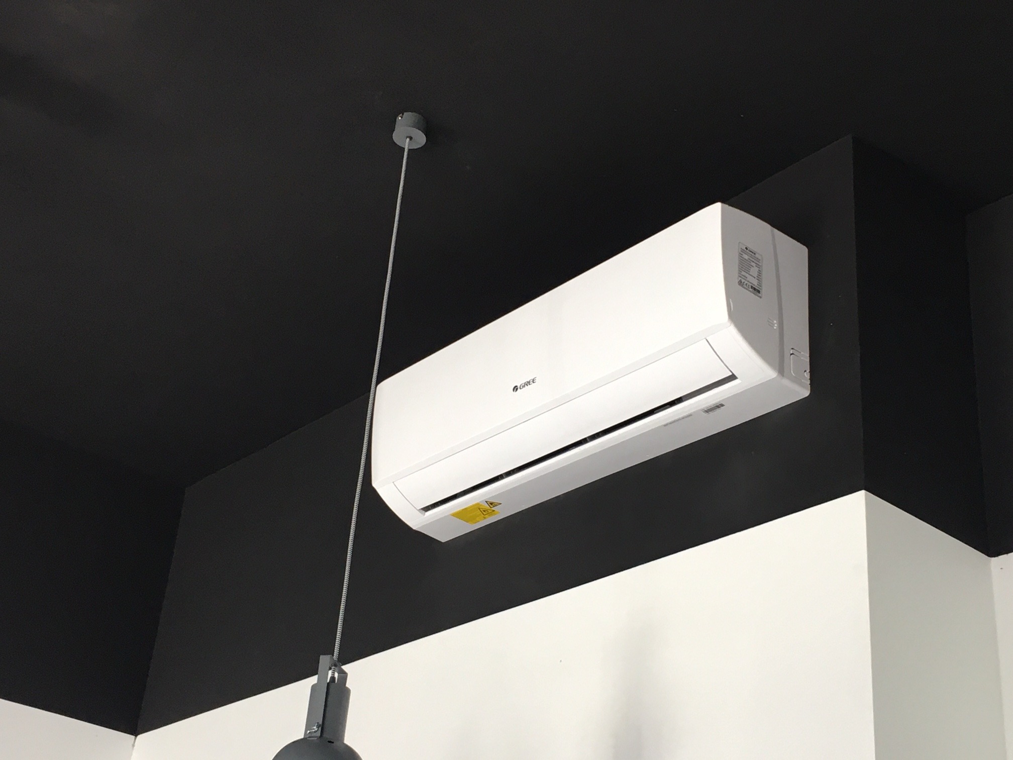 Biała jednostka wewnętrzna klimatyzacji marki Gree zamontowana na czarno-białej ścianie z lampą wiszącą