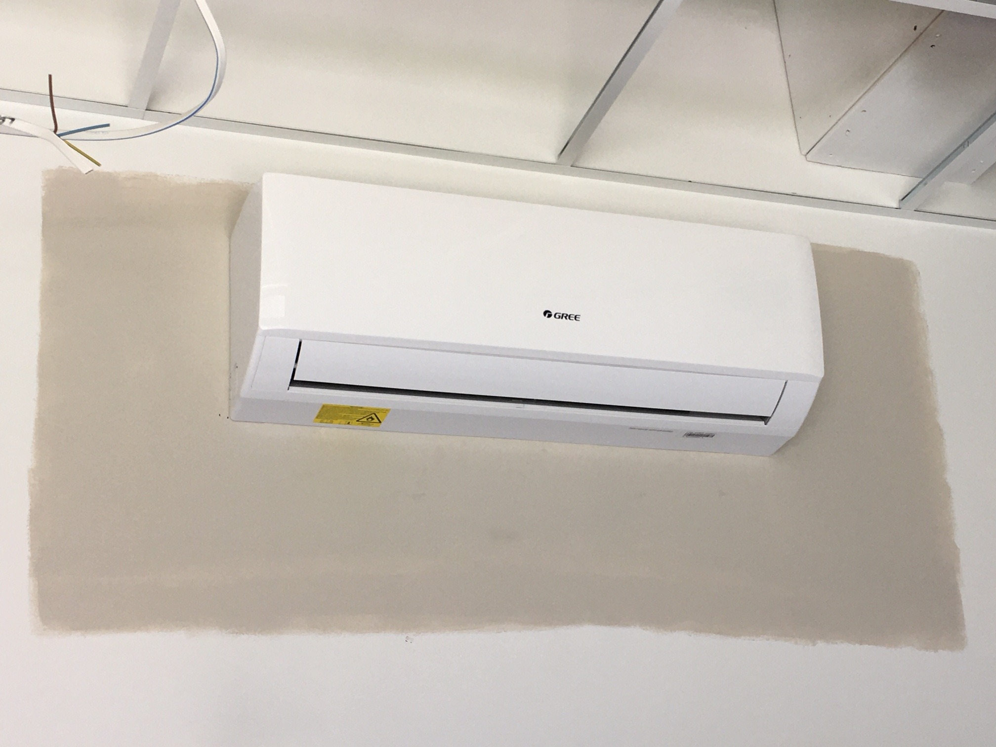 Biały klimatyzator marki Gree zamontowany na ścianie z widocznym niedomalowaniem farby, pod sufitem podwieszanym z widocznymi kablami.
