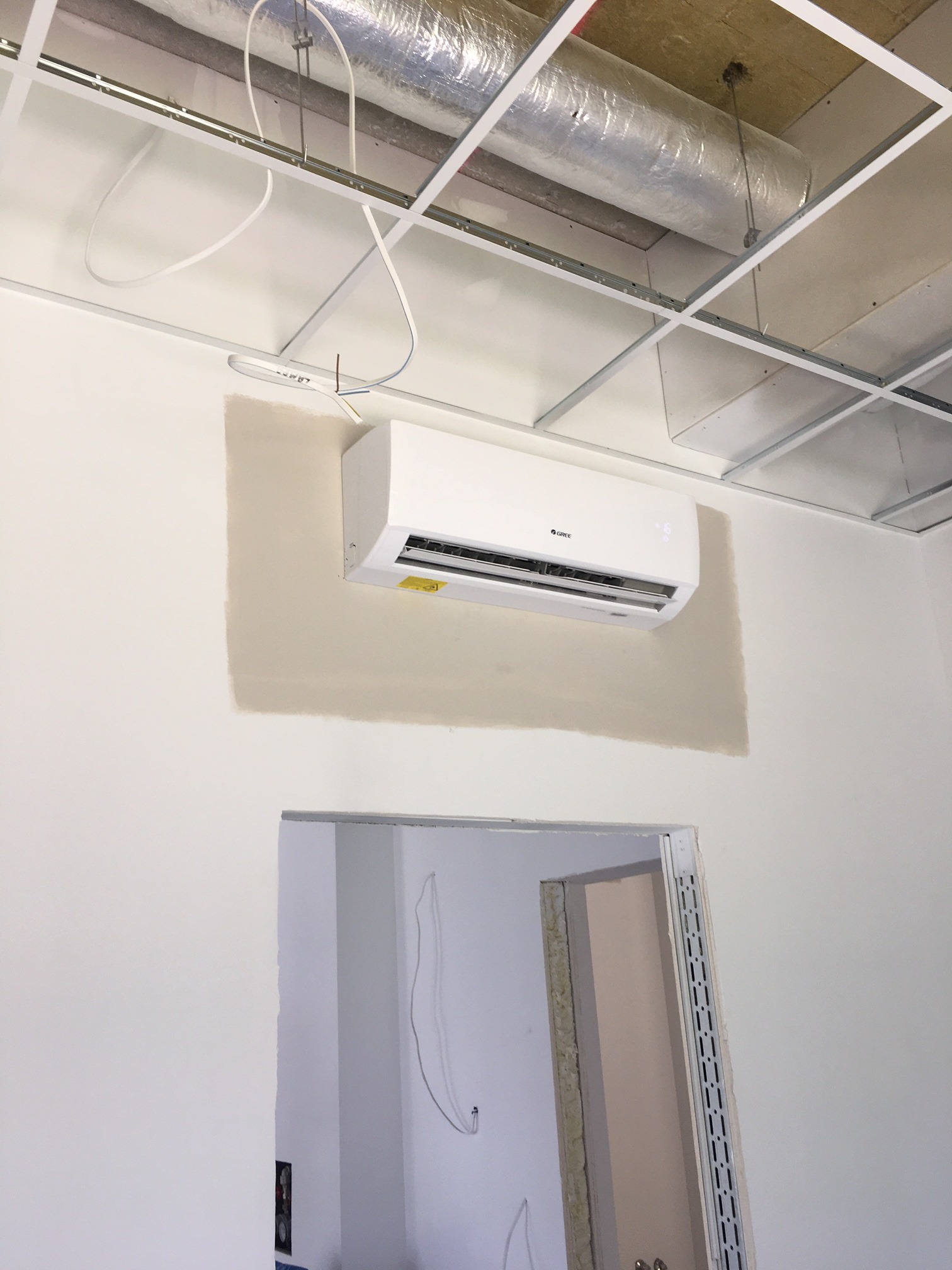 Biały klimatyzator marki Gree zamontowany na ścianie w pomieszczeniu biurowym w trakcie wykańczania, widoczne instalacje pod sufitem podwieszanym i niedokończone malowanie ściany.