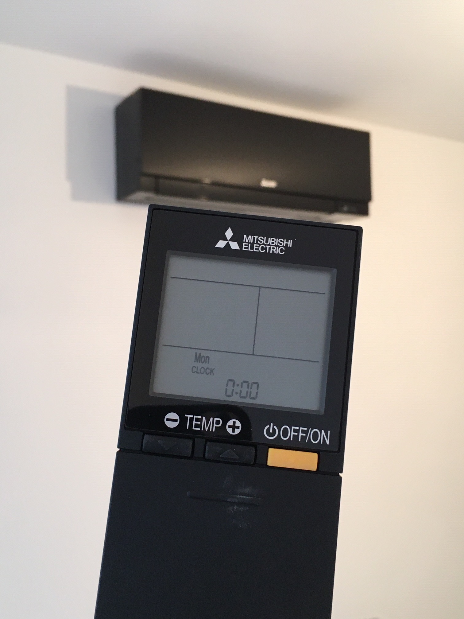 Czarny klimatyzator Mitsubishi Electric na ścianie, z bliska widoczny czarny pilot z wyświetlaczem pokazującym godzinę 0:00.