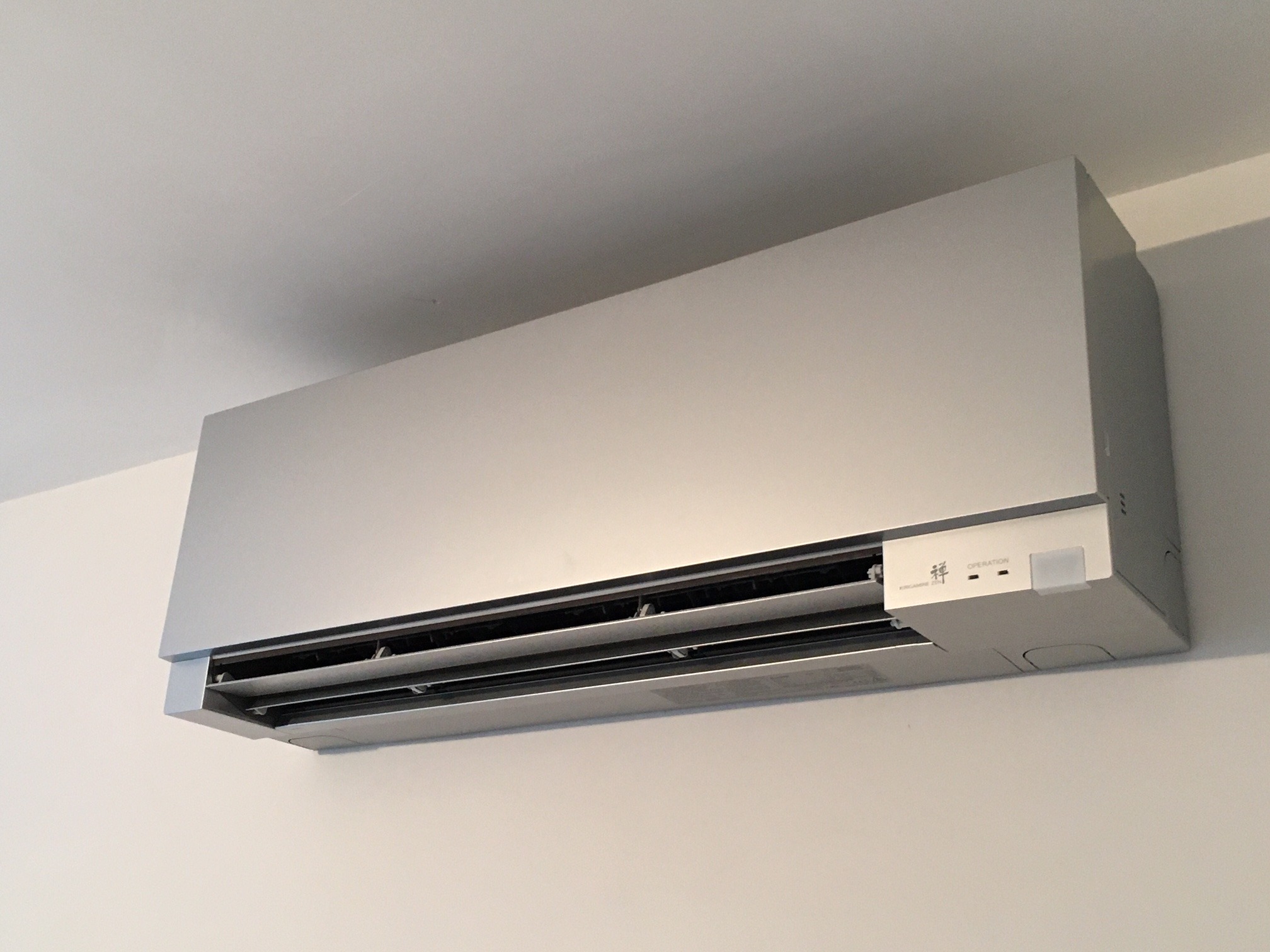 Nowoczesny, srebrny klimatyzator ścienny zamontowany blisko sufitu na białej ścianie, widoczne żaluzje i panel sterowania.